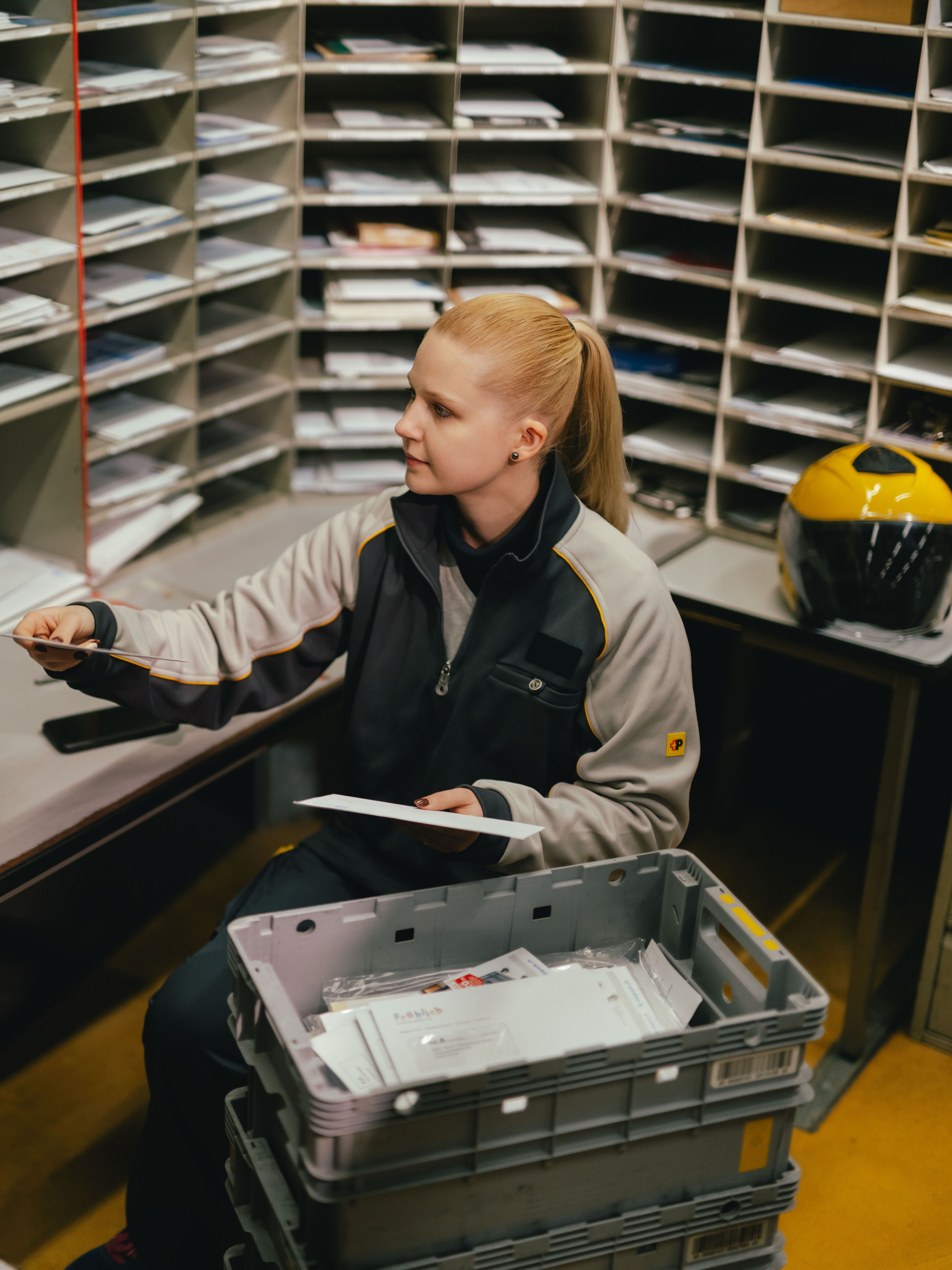 Eine Person sitzt in einem Raum mit vielen Postfächern und sortiert Briefe. Vor der Person steht eine graue Kunststoffkiste voller Sendungen. Auf dem Tisch liegen weitere Briefe, und neben dem Arbeitsplatz steht ein gelber Helm.