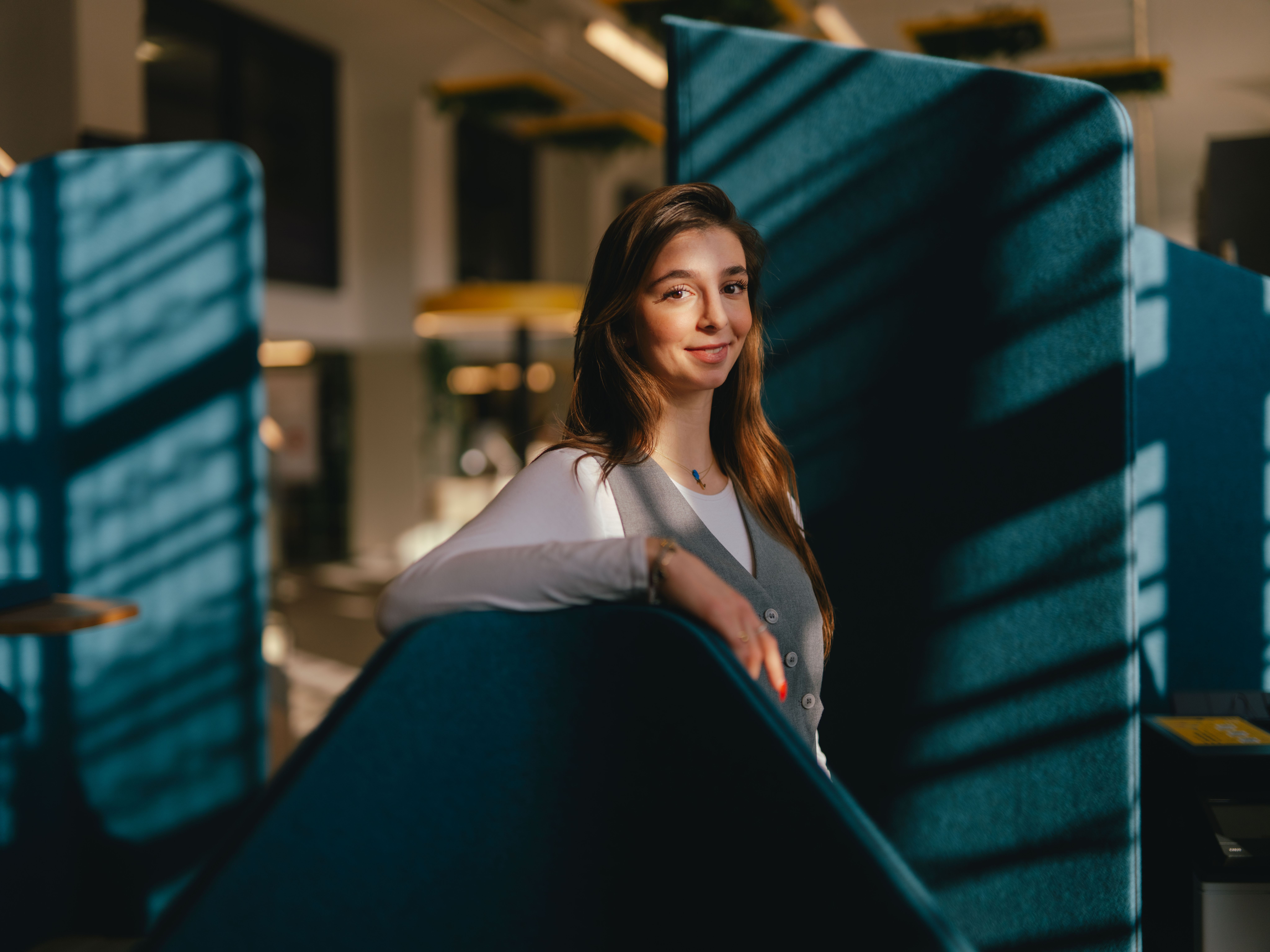 A person stands in a modern interior between tall, blue room dividers. Their body rotates slightly to the side and one arm rests loosely on the top edge of one of the room dividers. Strong sunlight falls through the large windows, casting clear shadow patterns on the blue areas.