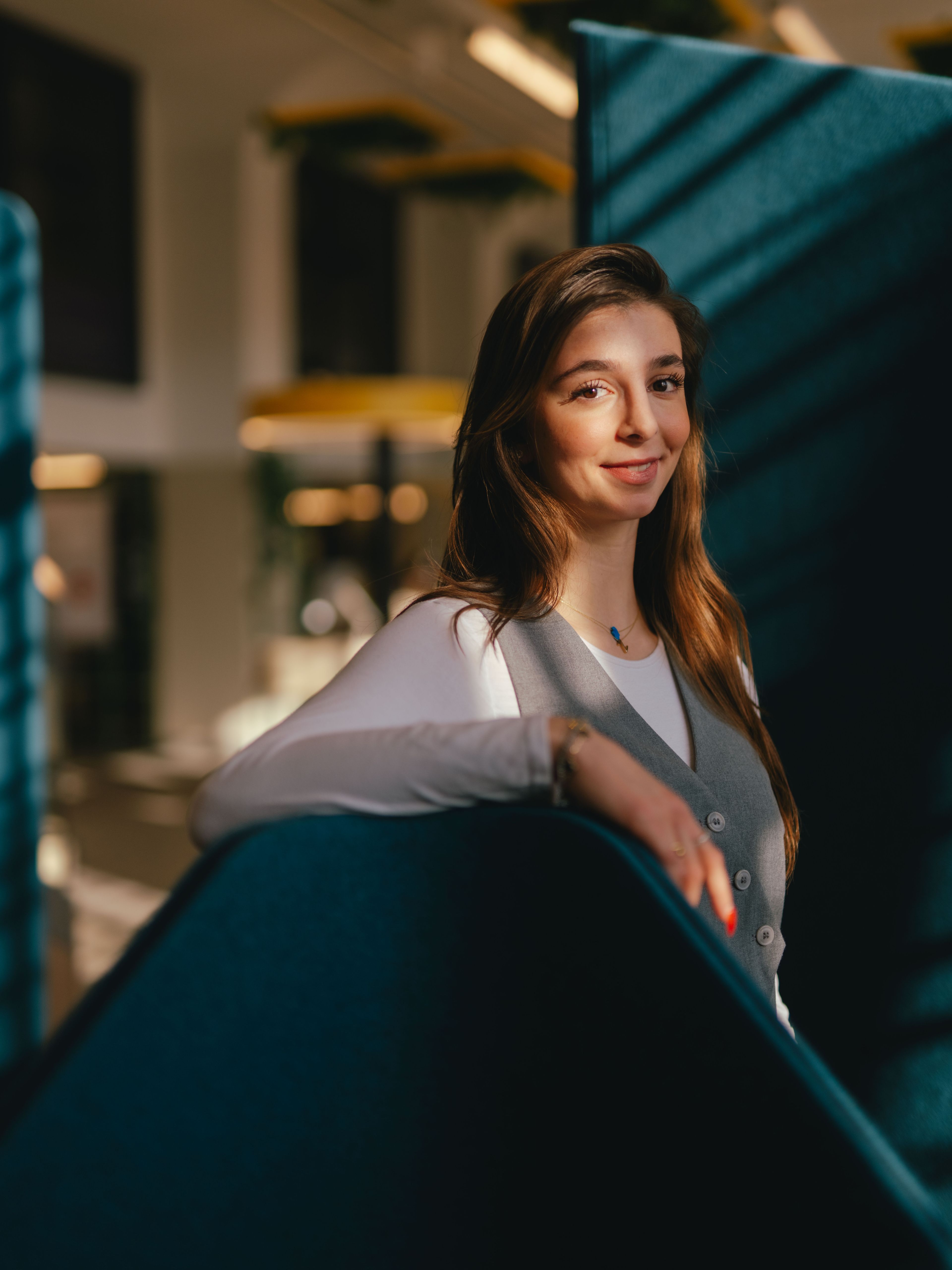 A person stands in a modern interior between tall, blue room dividers. Their body rotates slightly to the side and one arm rests loosely on the top edge of one of the room dividers. Strong sunlight falls through the large windows, casting clear shadow patterns on the blue areas.