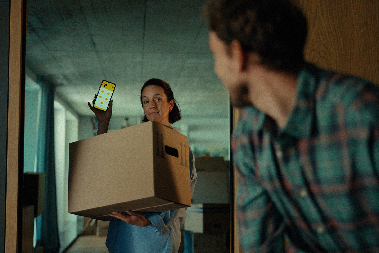 In einem Innenraum trägt eine Person einen grossen Umzugskarton in den Armen. In der anderen Hand hält die Person ein Smartphone hoch, dessen Display eine gelbe Benutzeroberfläche mit Symbolen zeigt. Im Vordergrund ist eine weitere Person seitlich erkennbar. Der Hintergrund umfasst Möbel, Kartons und einen Flur.