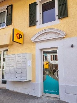 Entrée d’un bureau de poste dans un bâtiment jaune avec des volets gris. À gauche, une rangée de cases postales ; à droite, une porte vitrée avec le logo de la Poste et l’URL “mypostservice.ch”. Au-dessus de la porte, un arc décoratif blanc.