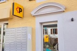 Entrée d’un bureau de poste dans un bâtiment jaune avec des volets gris. À gauche, une rangée de cases postales ; à droite, une porte vitrée avec le logo de la Poste et l’URL “mypostservice.ch”. Au-dessus de la porte, un arc décoratif blanc.