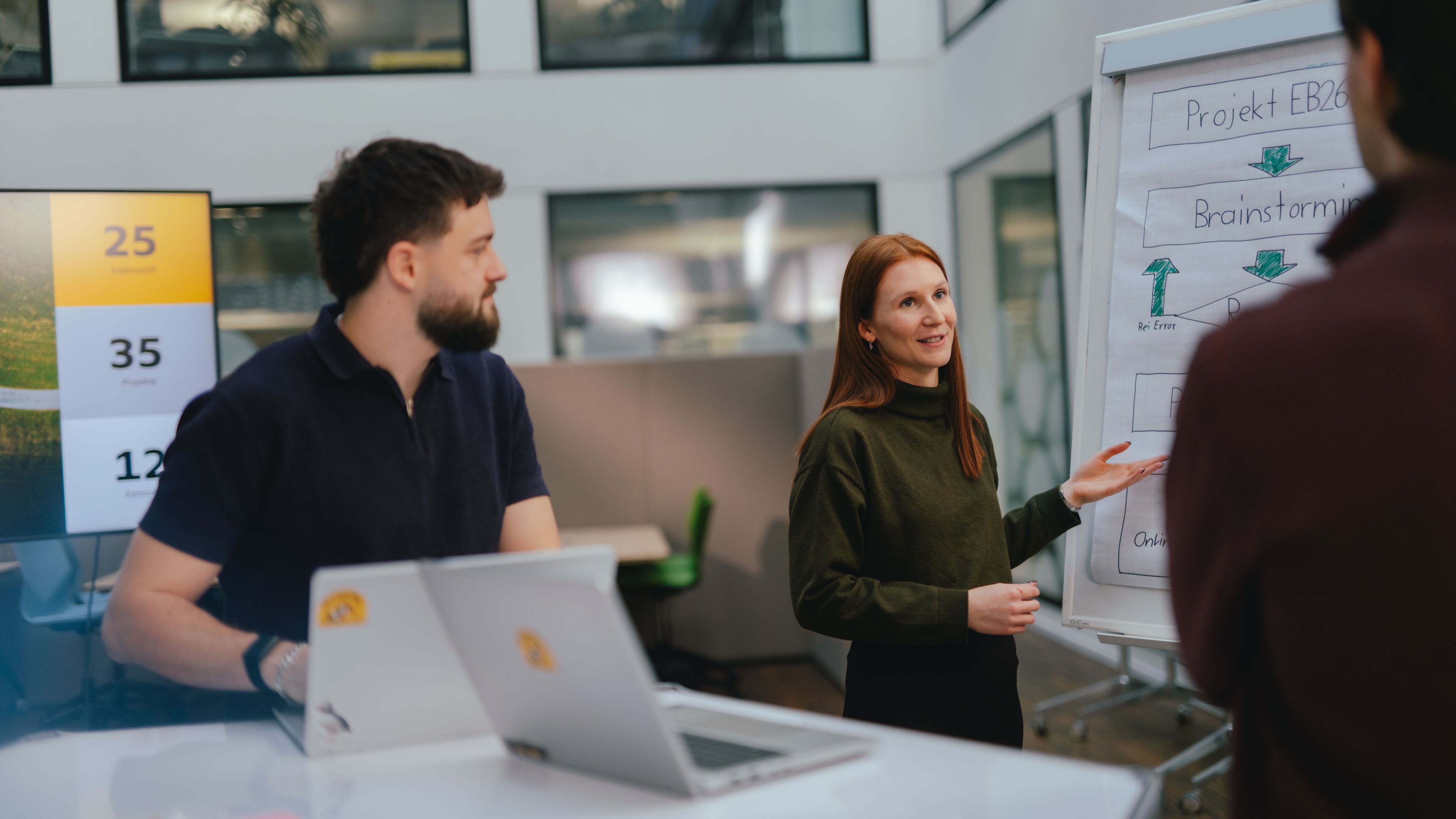 Plusieurs personnes se trouvent dans un espace de bureaux moderne. Une personne montre un tableau chevalet sur lequel figurent des points clés et des flèches écrits à la main. Une autre personne se tient à côté et regarde en direction du tableau chevalet. Au premier plan, un ordinateur portable déplié est visible sur une table. La salle est dotée de grandes fenêtres, de parois vitrées et d’une architecture intérieure claire.