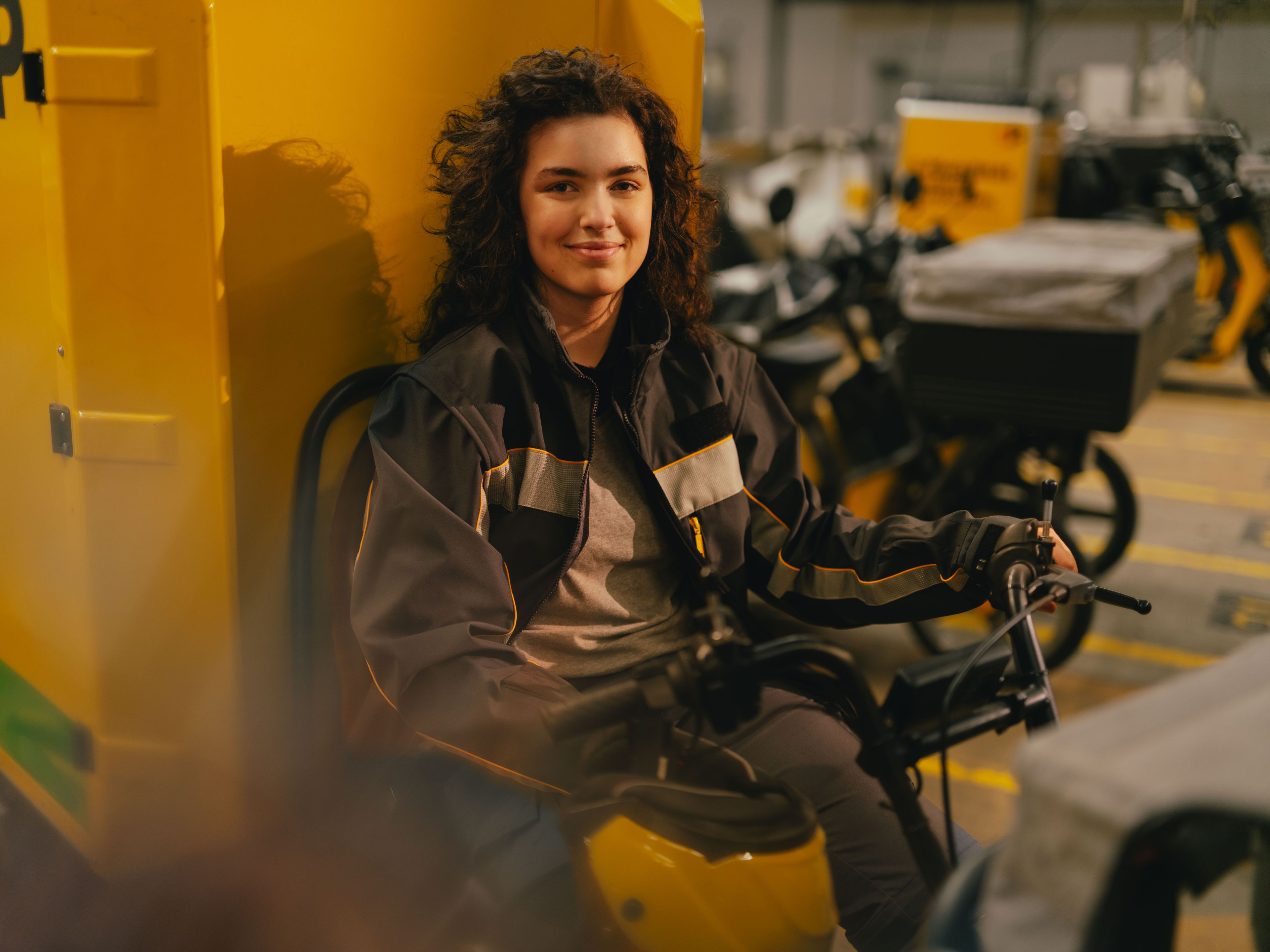 Eine Person sitzt in einer grossen Halle auf einem gelben Fahrzeug, das wie ein elektrisches Transport- oder Zustellfahrzeug wirkt. Die Person trägt eine dunkel gehaltene Arbeitsjacke mit hellen Streifen. Im Hintergrund stehen mehrere weitere Fahrzeuge dicht nebeneinander in einer markierten Parkzone.