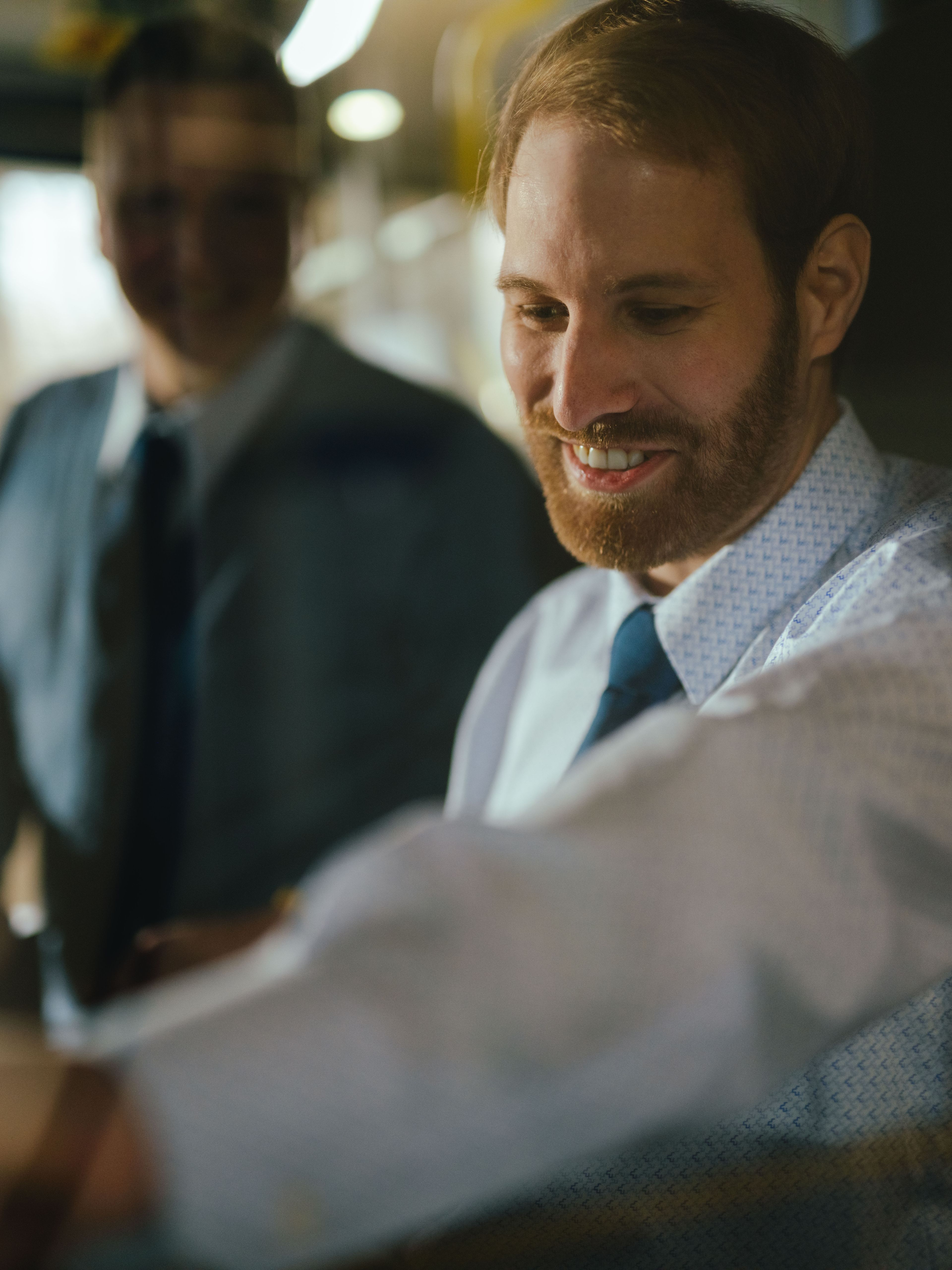 Two people are in the driver’s area of a vehicle. One person is seated at the wheel and reaching for a control, while the other stands beside them. The steering wheel, dashboard controls, and interior lighting are visible.