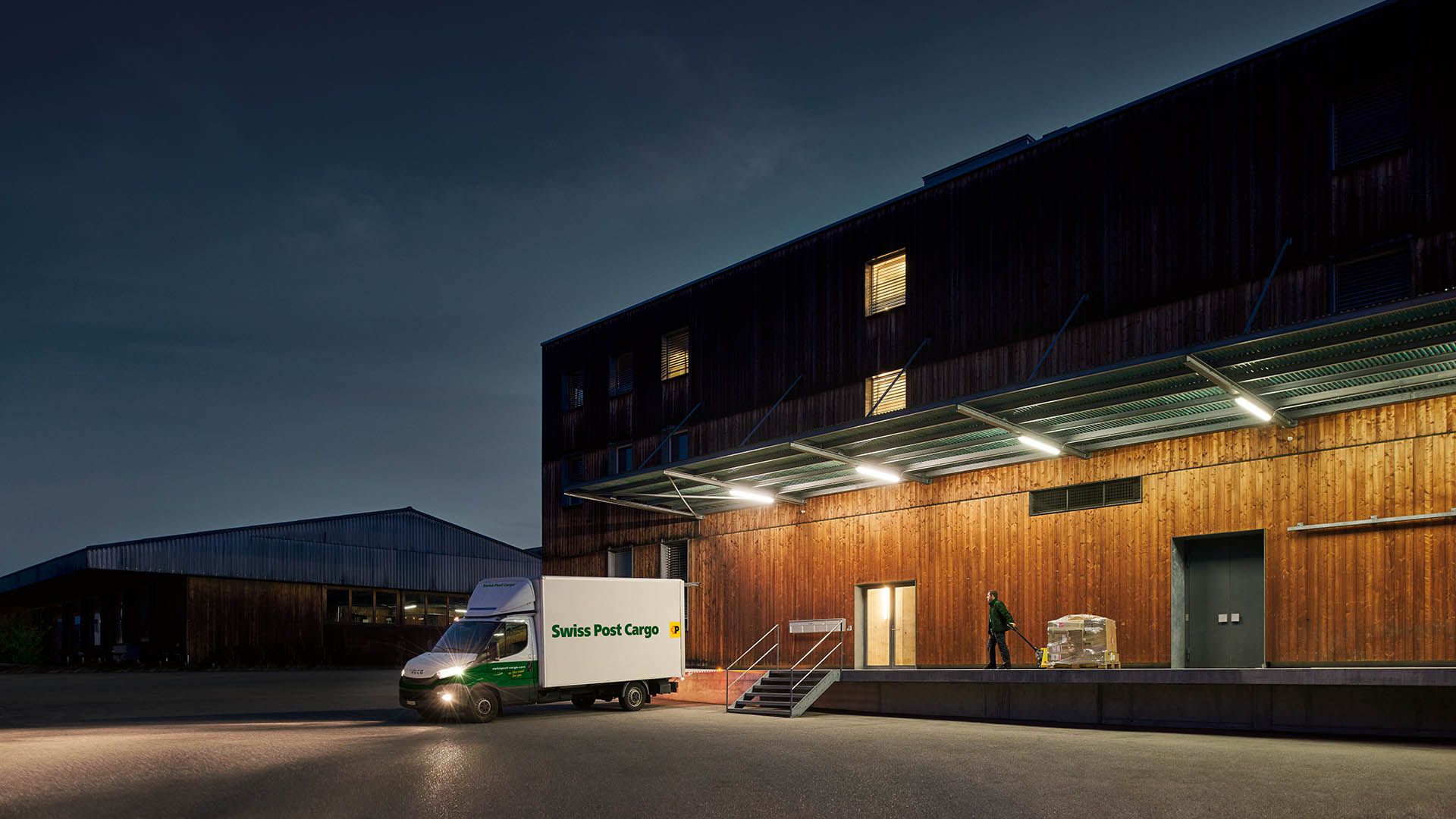 A night courier from Swiss Post Cargo delivers goods to a warehouse building before sunrise.