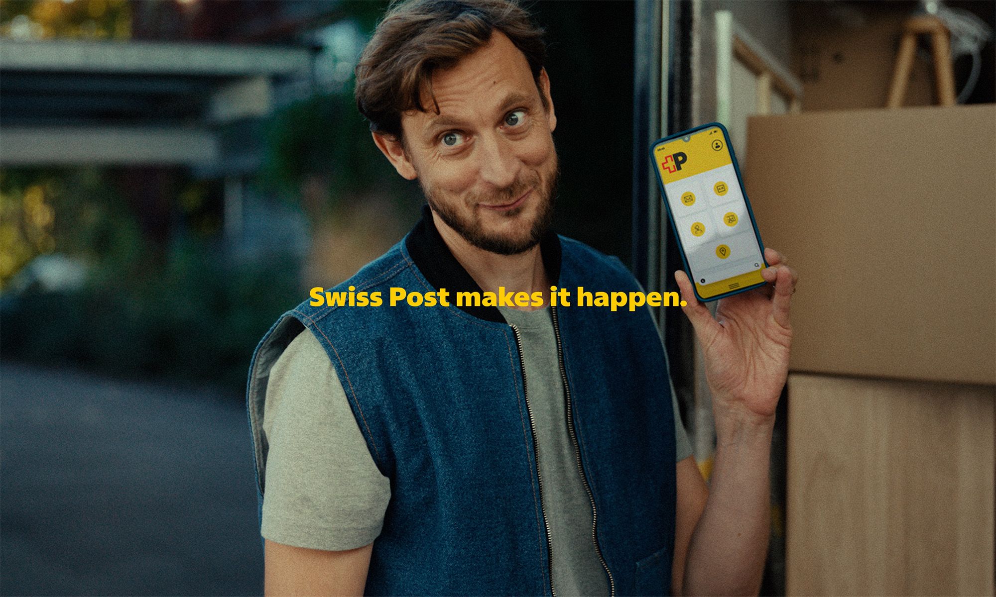 A person is standing in front of an open transport vehicle and holding up a smartphone displaying a yellow interface with several icons. In front of the person are moving boxes and wooden surfaces. In the background, trees, a building, and a covered structure are visible. The yellow text “Die Post macht’s möglich.” appears in the center of the image.