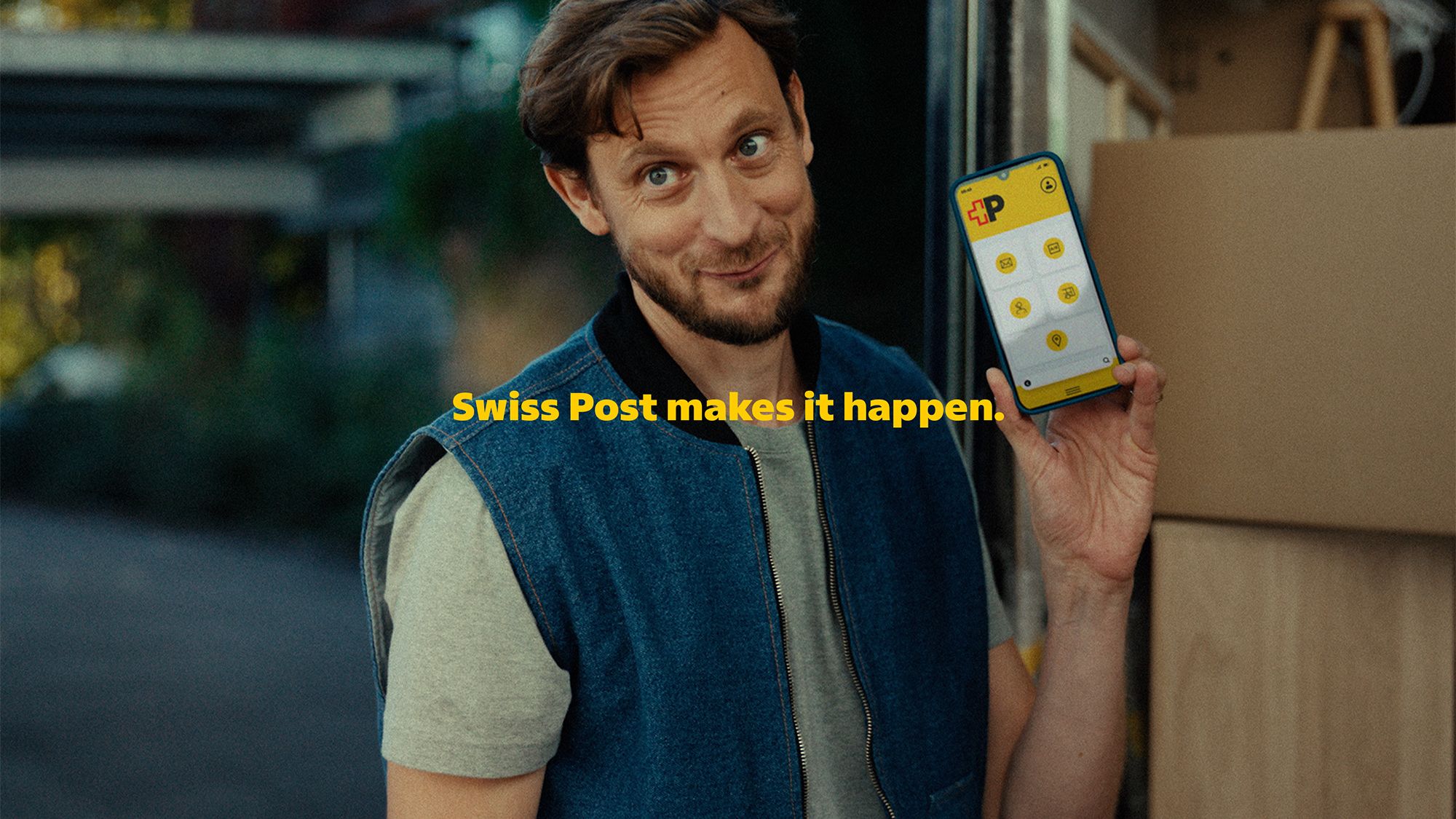 A person is standing in front of an open transport vehicle and holding up a smartphone displaying a yellow interface with several icons. In front of the person are moving boxes and wooden surfaces. In the background, trees, a building, and a covered structure are visible. The yellow text “Die Post macht’s möglich.” appears in the center of the image.