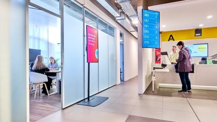 Interno di una filiale postale moderna. A sinistra uffici consulenza con pareti in vetro, due persone sedute a un tavolo. A destra un banco con una persona. Schermi digitali mostrano numeri e indicazioni.