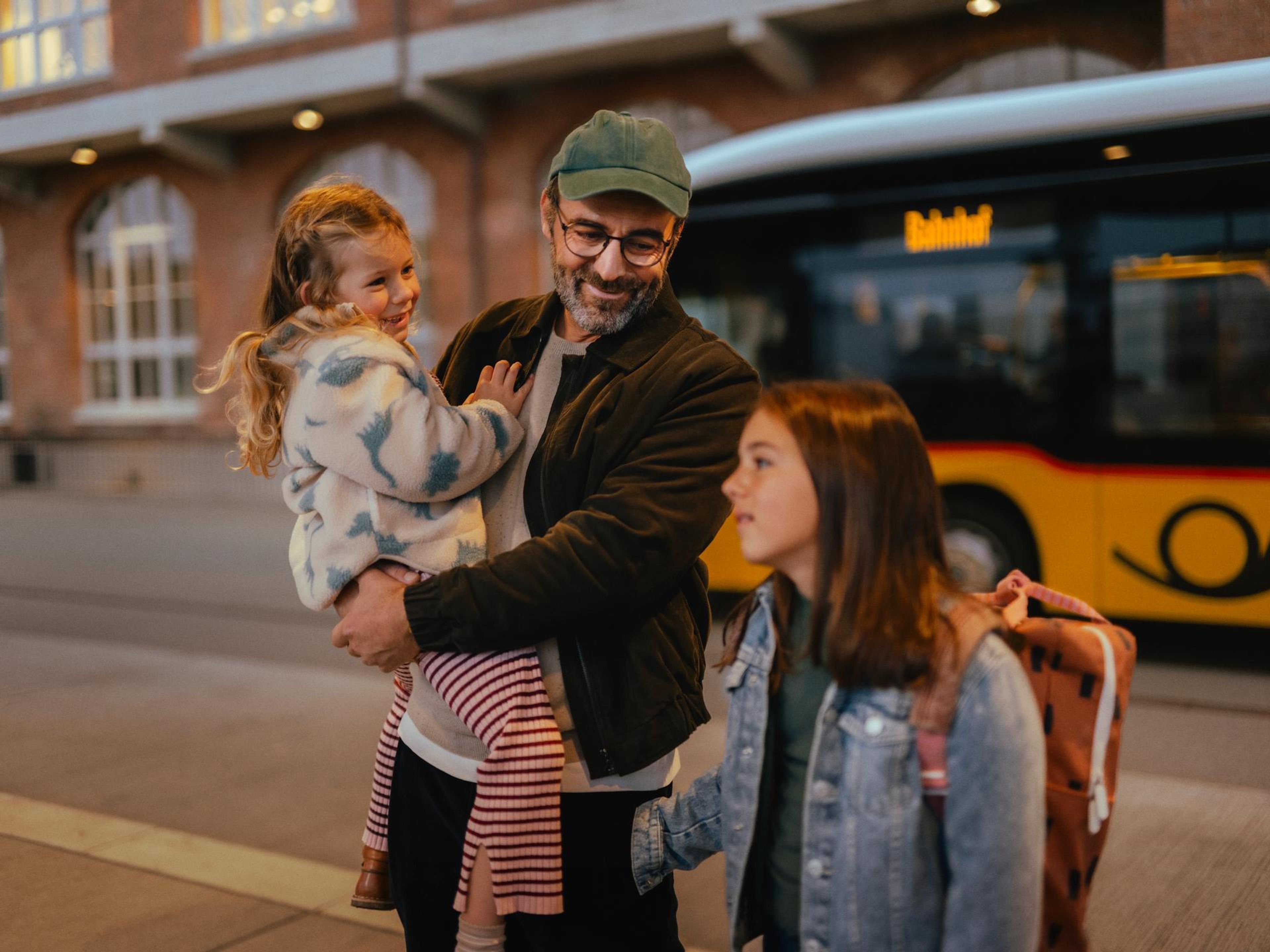 Trois personnes se tiennent à un arrêt de bus devant un bus jaune. Une personne porte un enfant dans les bras, tandis qu’une autre personne avec un sac à dos se tient à côté. En arrière‑plan, on voit un bâtiment en briques avec des fenêtres éclairées.