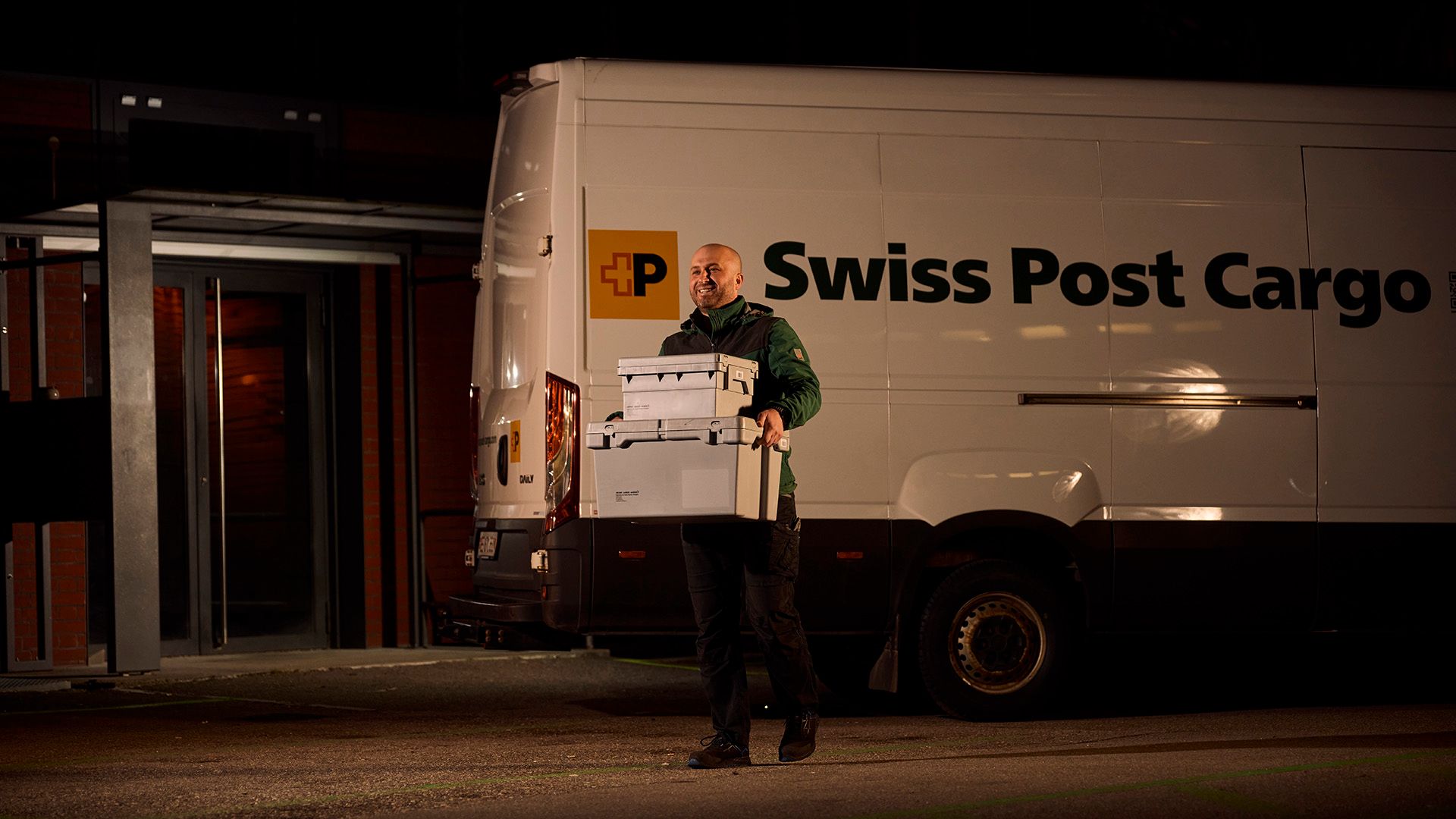 Swiss Post Cargo employees with parcels in front of the delivery vehicle at night.