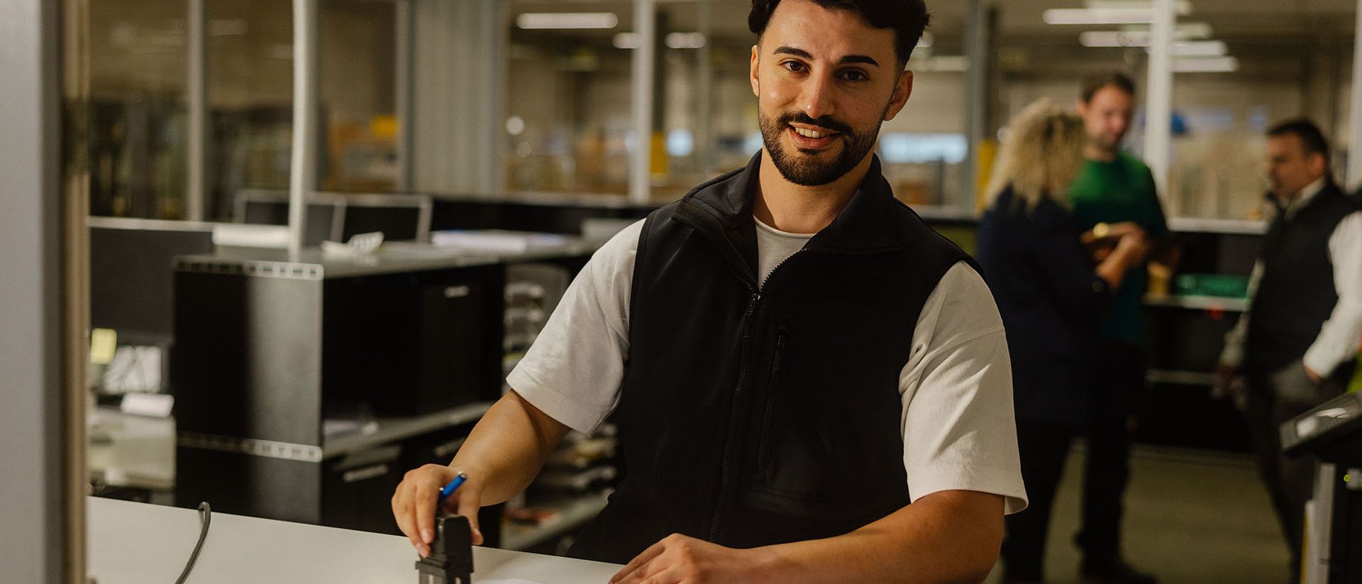 A customs officer stamps documents at a counter, with an office with several people in the background.