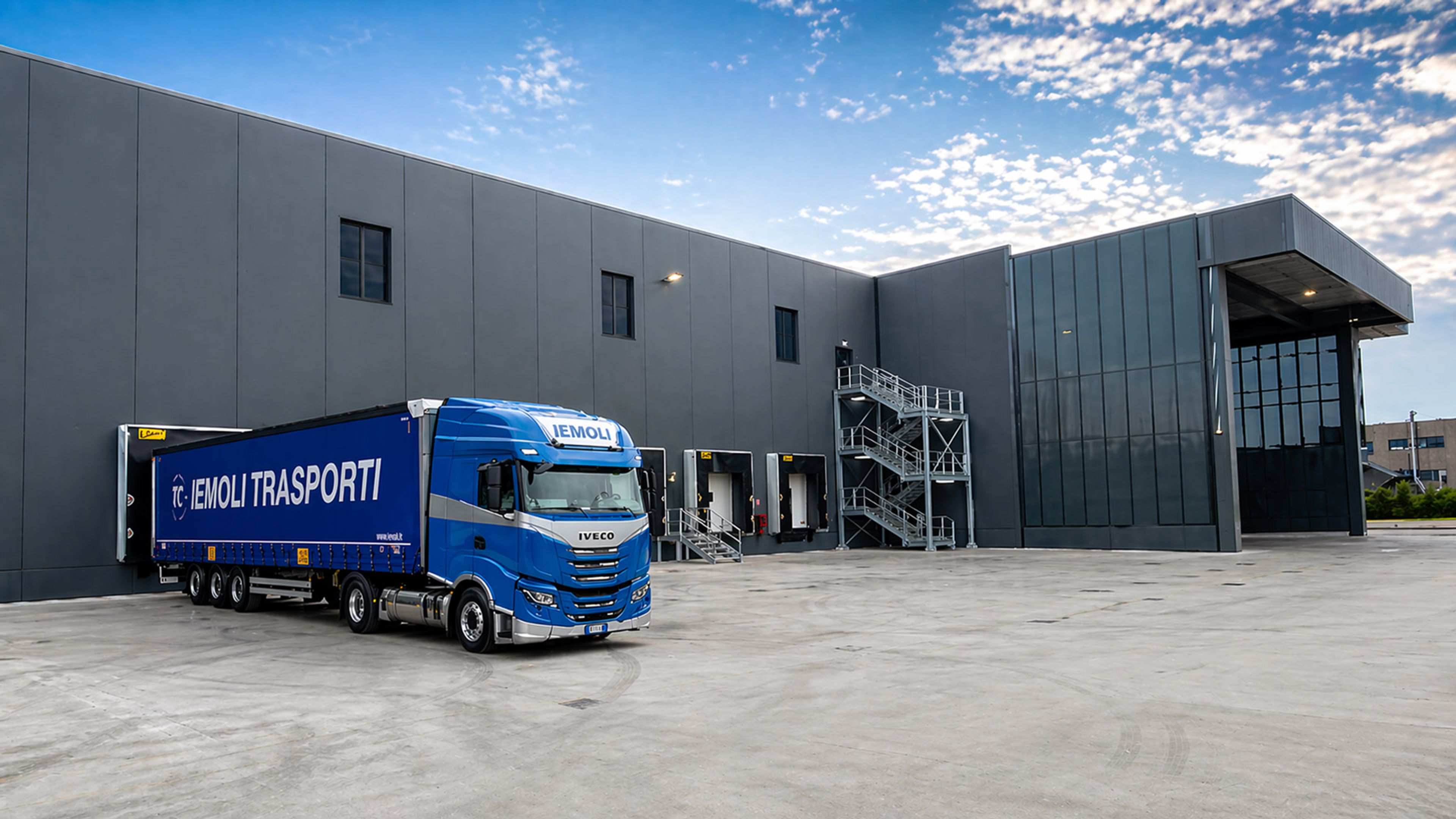 Autoarticolato Iemoli blu davanti a un moderno centro logistico con diverse piattaforme di caricamento e un capannone industriale grigio.