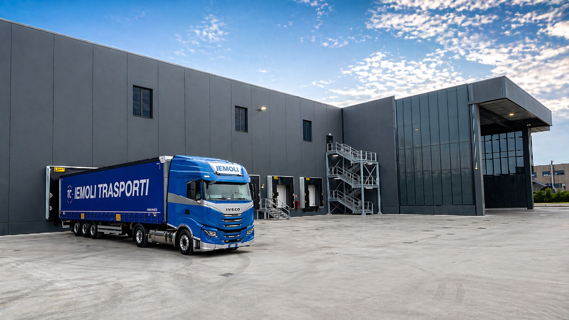 Autoarticolato Iemoli blu davanti a un moderno centro logistico con diverse piattaforme di caricamento e un capannone industriale grigio.
