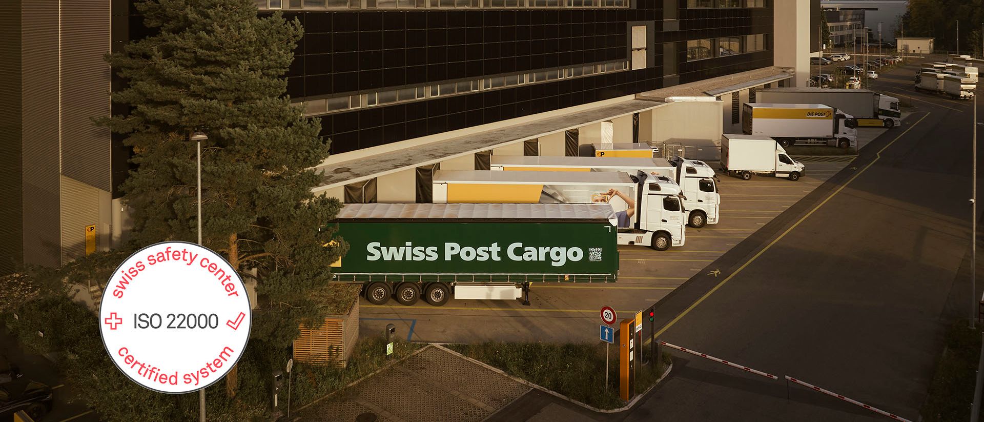 Swiss Post Cargo’s modern Villmergen logistics center with loading ramps and several trucks loading and unloading goods, supplemented by an ISO 22000 certificate label.