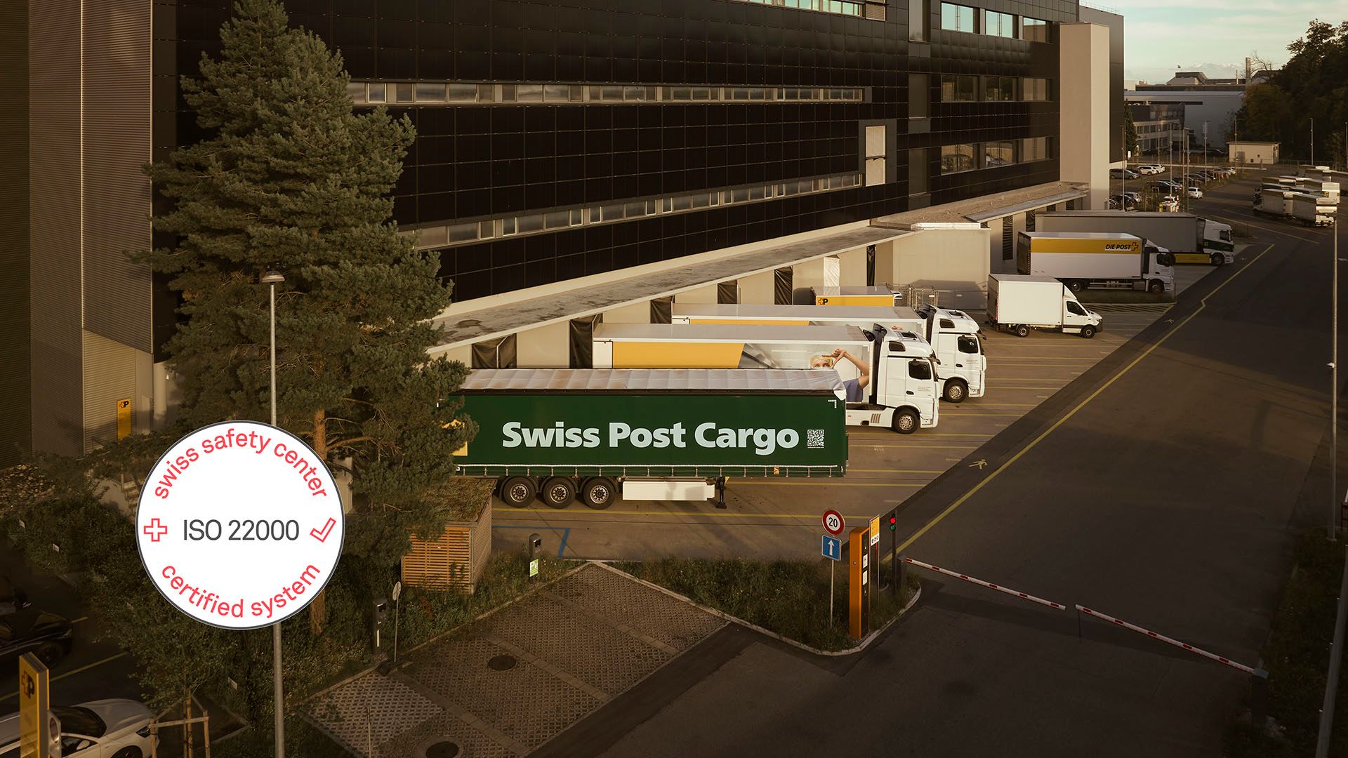 Swiss Post Cargo’s modern Villmergen logistics center with loading ramps and several trucks loading and unloading goods, supplemented by an ISO 22000 certificate label.