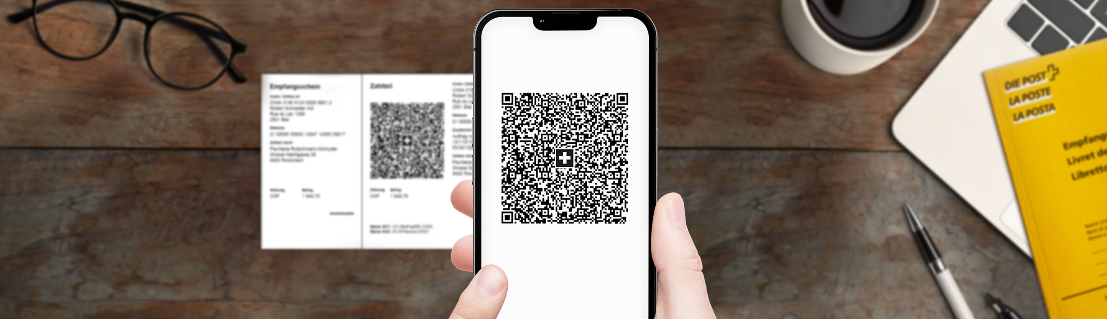 Mano che tiene uno smartphone con un codice QR sullo schermo. Sul tavolo ci sono una fattura QR, occhiali, un libretto giallo della Posta, un portatile, una penna e una tazza di caffè.