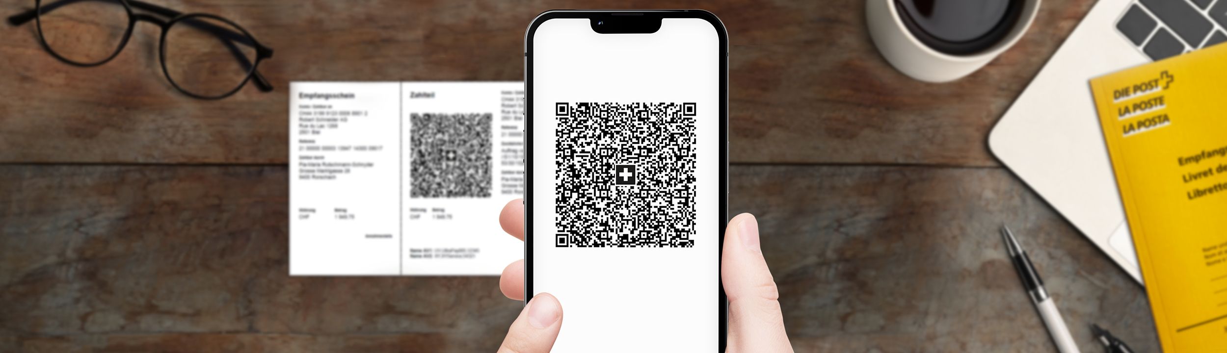 Mano che tiene uno smartphone con un codice QR sullo schermo. Sul tavolo ci sono una fattura QR, occhiali, un libretto giallo della Posta, un portatile, una penna e una tazza di caffè.