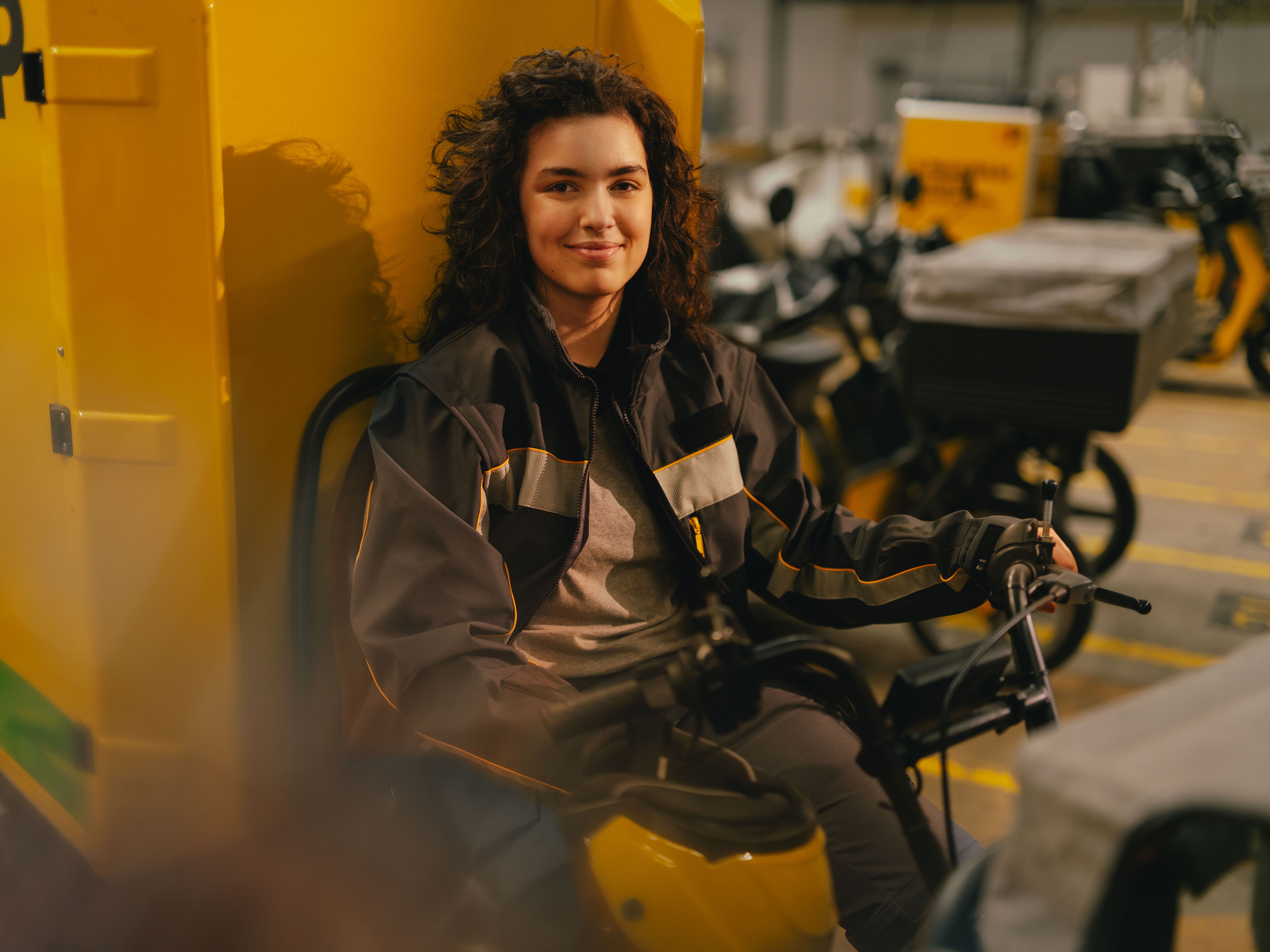 Une personne est assise dans un grand hall sur un véhicule jaune ressemblant à un engin de transport ou de livraison électrique. La personne porte une veste de travail sombre avec des bandes claires. À l’arrière‑plan, plusieurs autres véhicules sont alignés dans une zone de stationnement marquée.