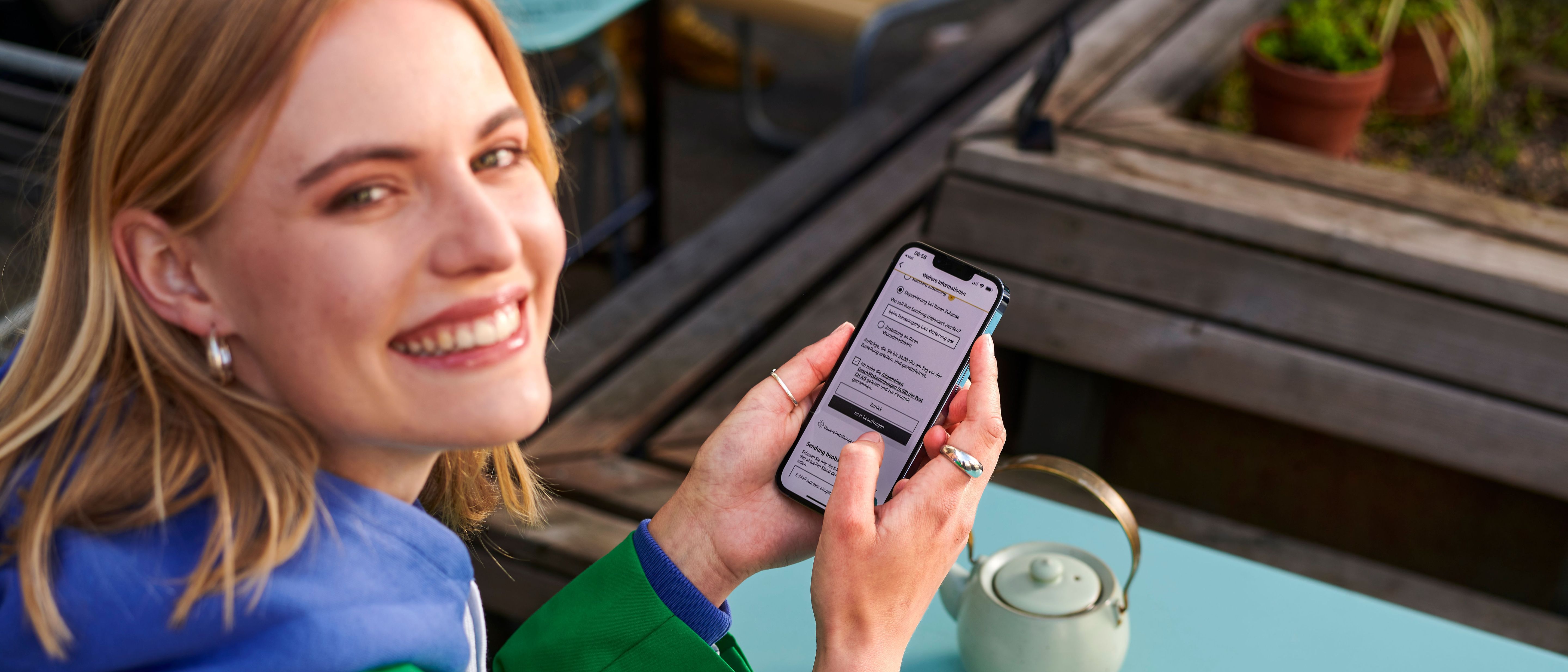 Person sitzt an einem Tisch im Freien und hält ein Smartphone in der Hand. Auf dem Tisch steht eine helle Teekanne. Die Person betrachtet eine geöffnete App oder Webseite auf dem Bildschirm.