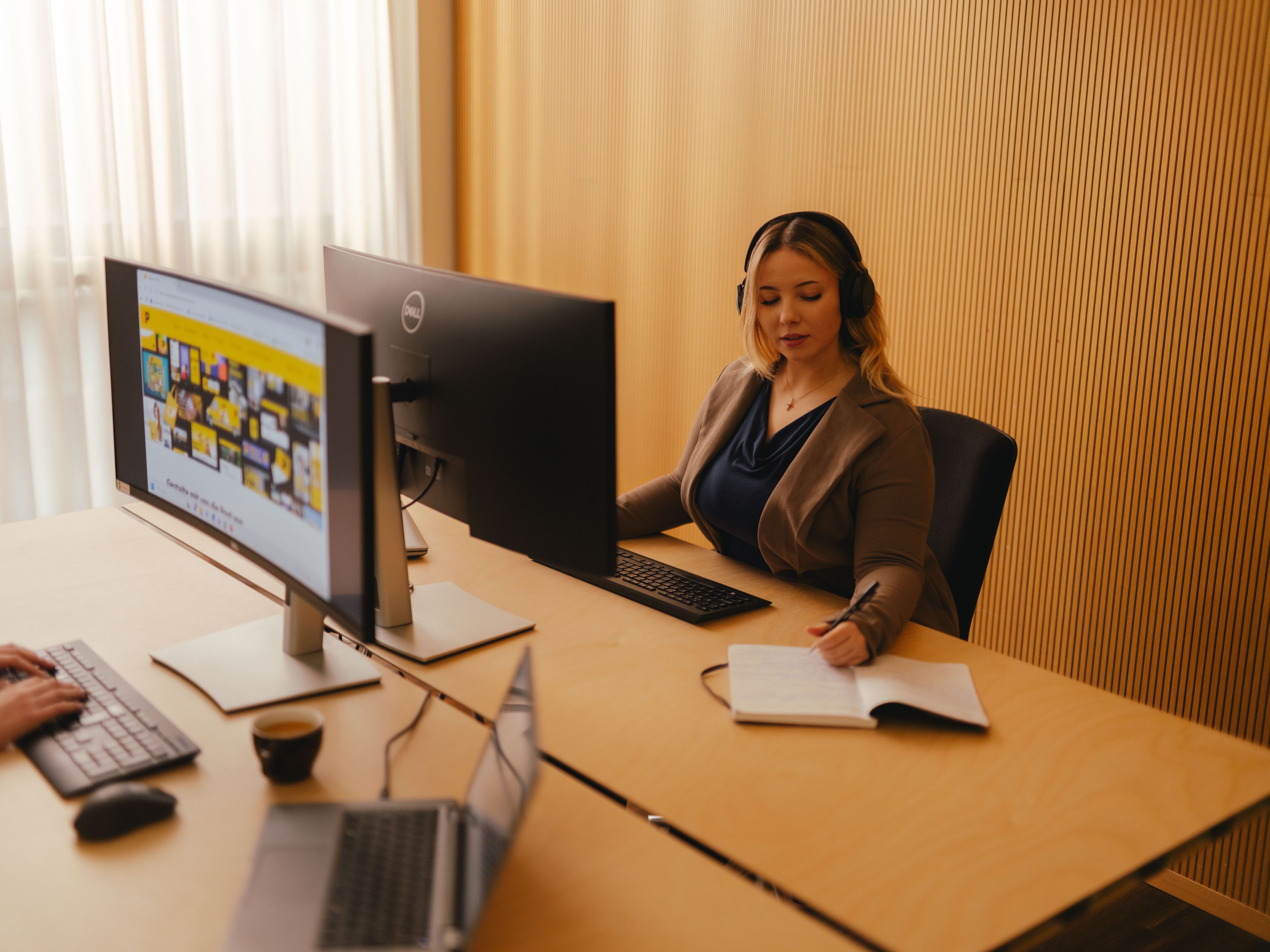A person sits at a desk in an office environment, working with a large monitor and a keyboard. The person is wearing a headset and has an open notebook in front of them. In the foreground, another laptop is placed on a second desk. The room features light curtains and a wall with a vertical pattern.