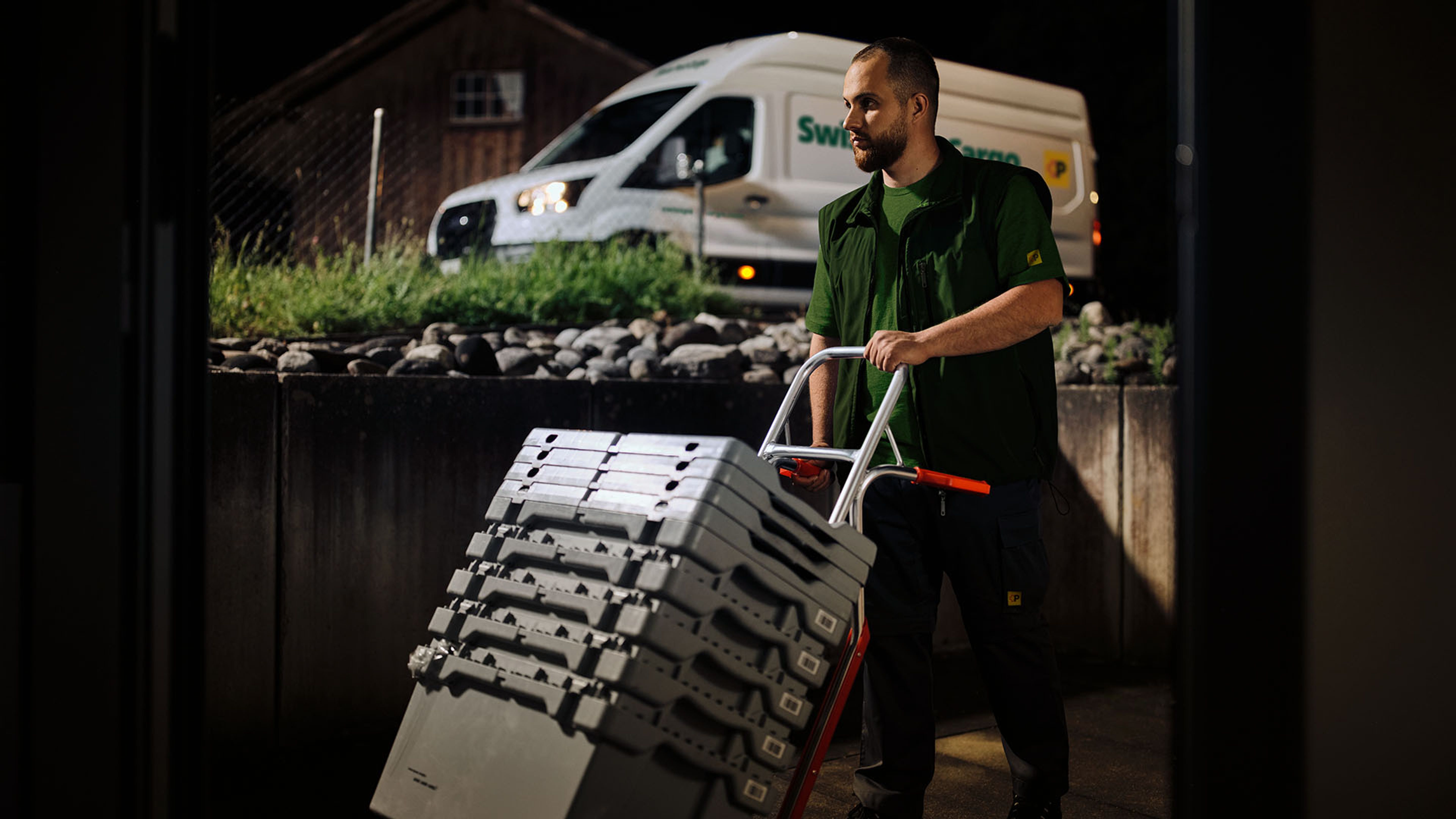 A Swiss Post Cargo night courier delivers medical goods with a hand trolley, with a delivery vehicle in the background.