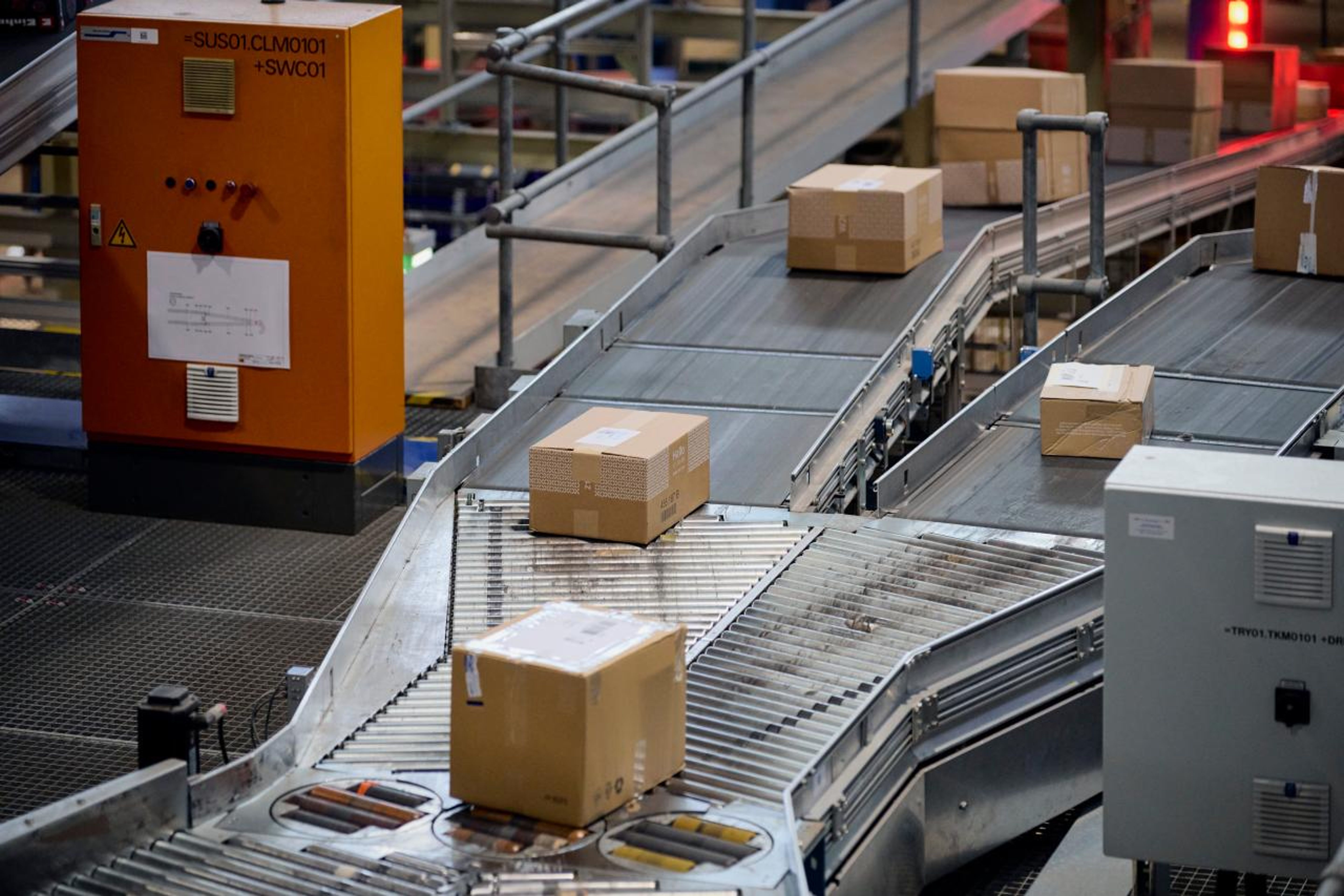 An automated conveyor system transports several parcels on taxiways in a logistics center. An orange control element is visible, which indicates an industrial environment. The scene focuses on the efficient movement of consignments.