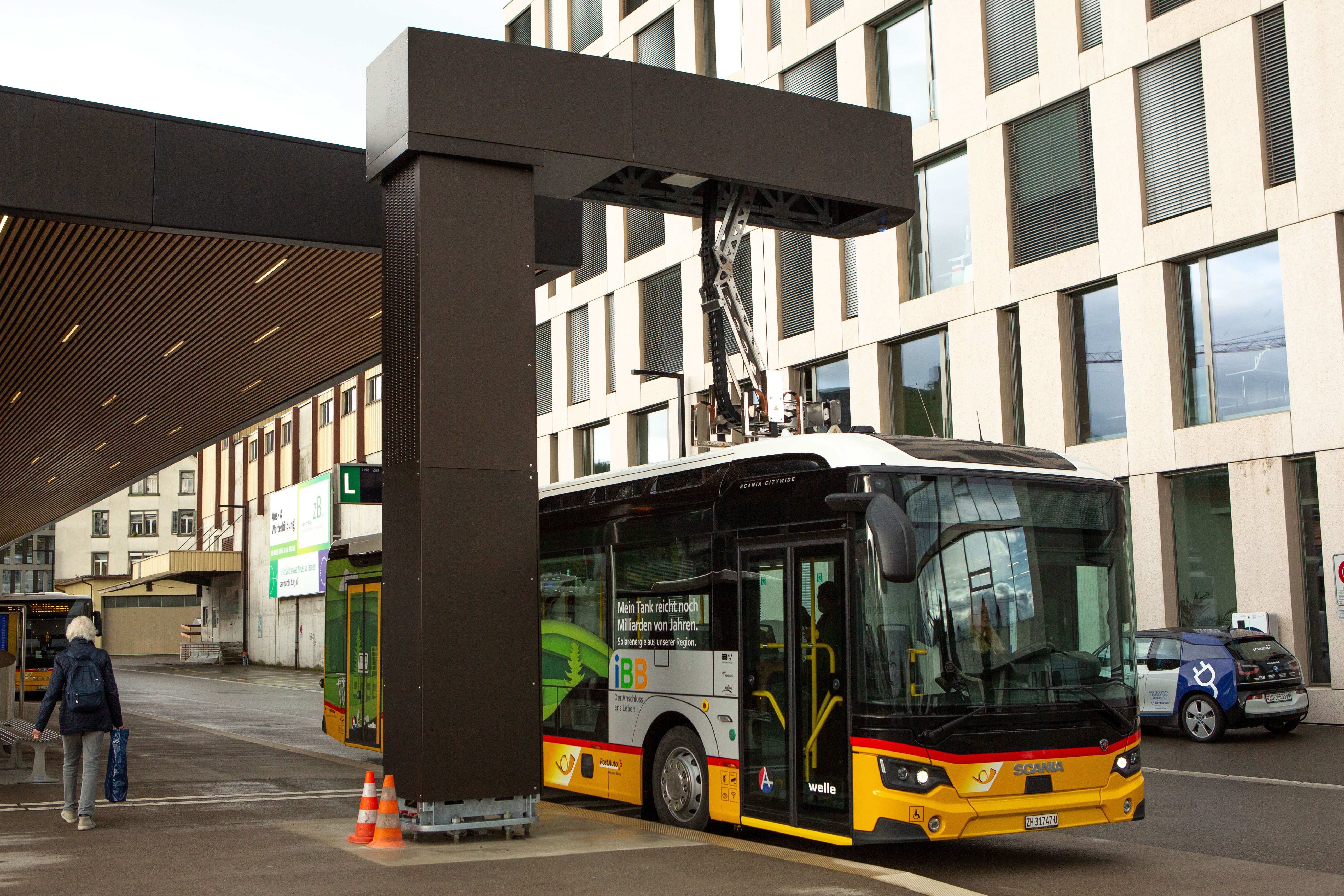 Gelb-schwarzer Elektrobus an Ladestation vor Gebäude, Person mit Rucksack geht vorbei