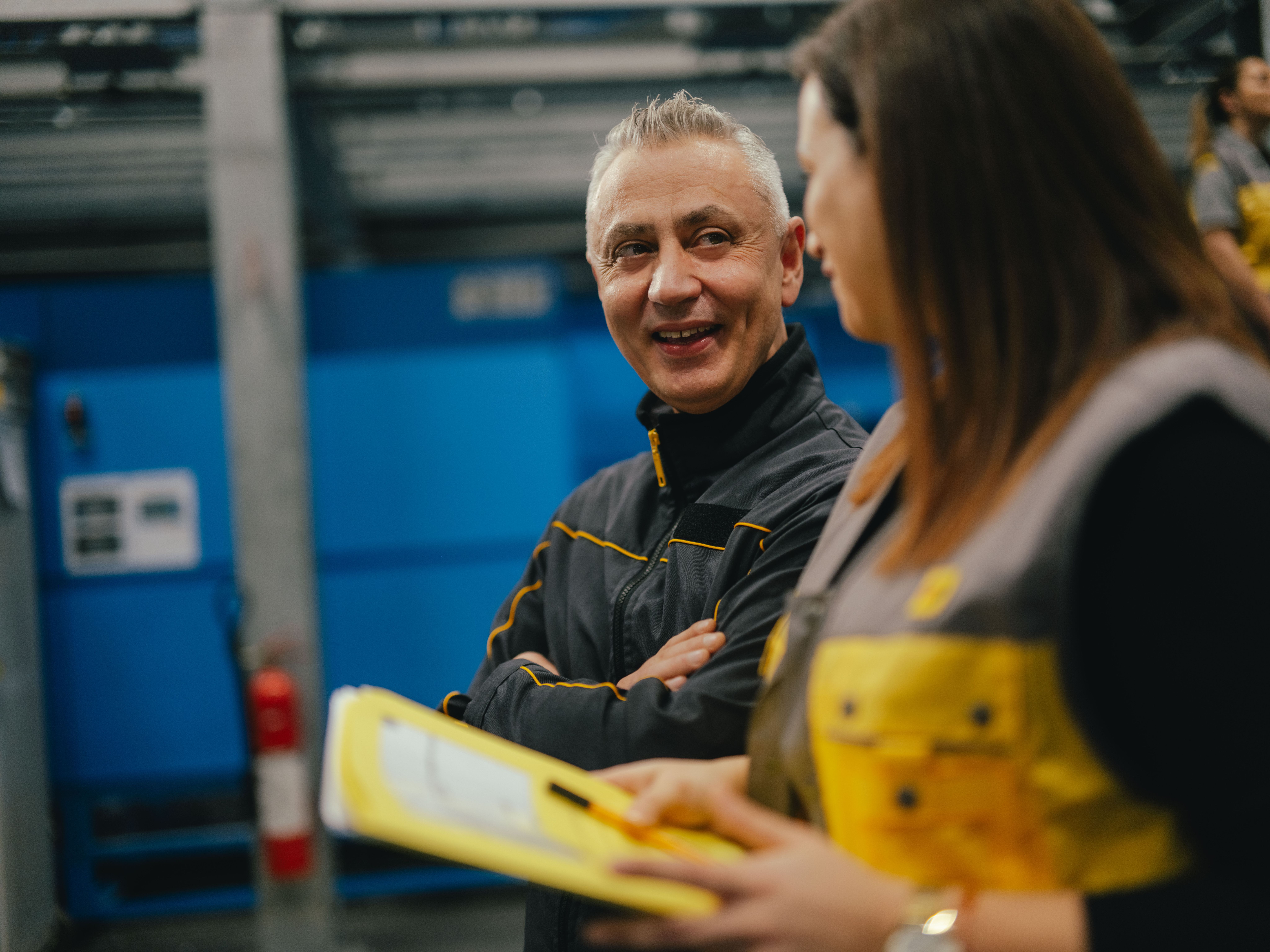 Two people are in an industrial setting, looking together at a yellow clipboard held by one of them. Both are wearing dark work clothing with yellow accents. In the background, blue machine housings, metal structures, and a fire safety station are visible.