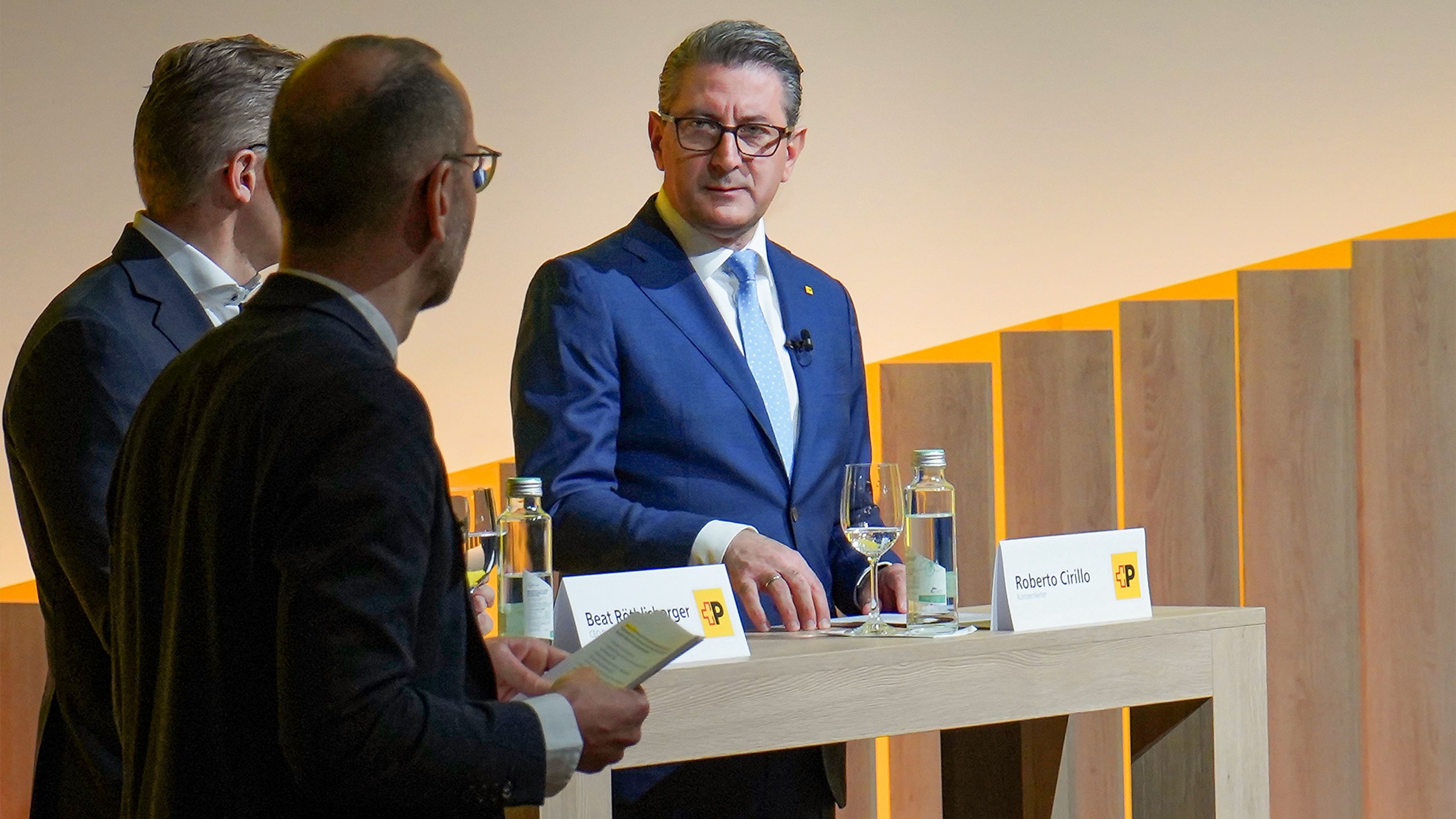 Ein Mann in Anzug steht an einem Pult mit Wasserflaschen und Namensschildern.