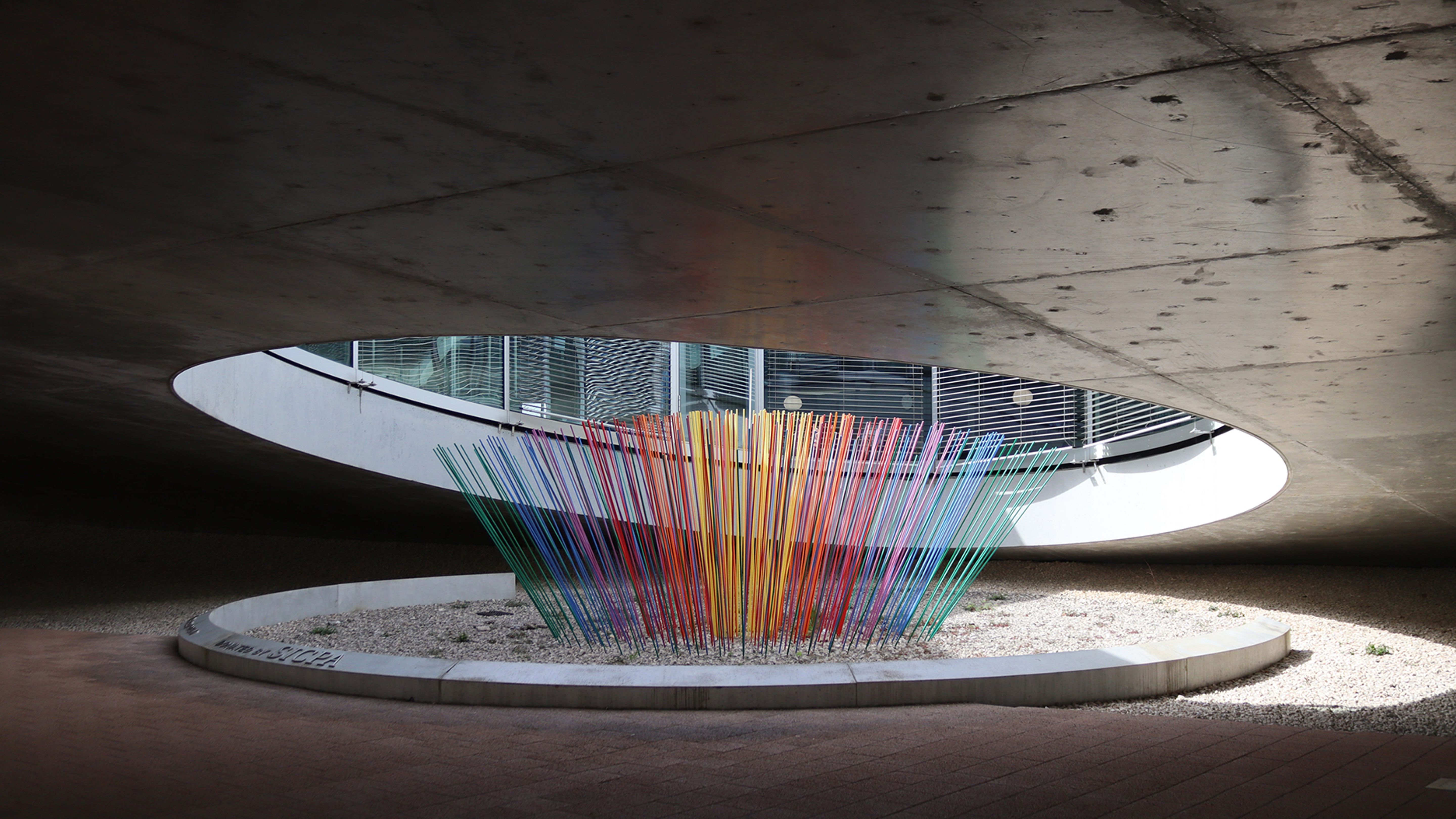 Une œuvre d’art multicolore composée de tiges fines se dresse sous une toiture en béton.