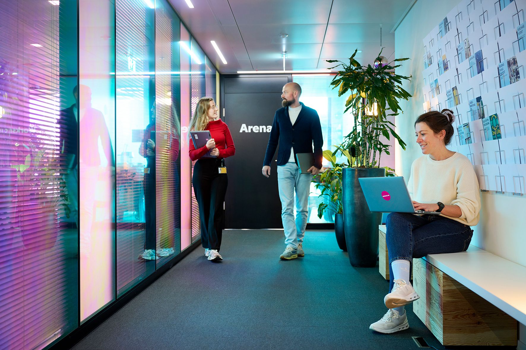 Personen in einem modernen Bürogebäude, zwei gehend, eine sitzend mit Laptop.