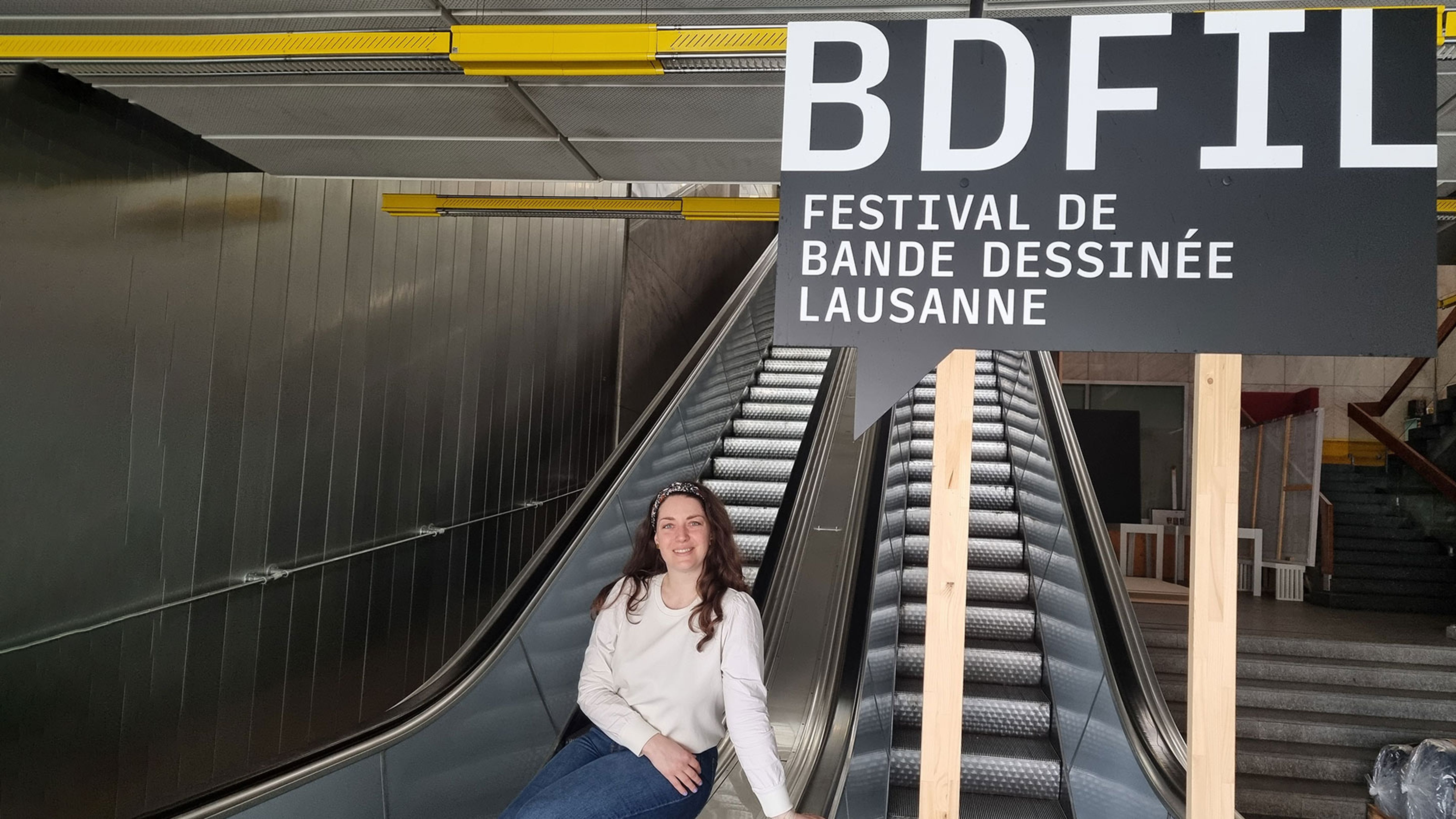 Una persona siede su una scala mobile davanti a un cartello per un festival a Losanna.