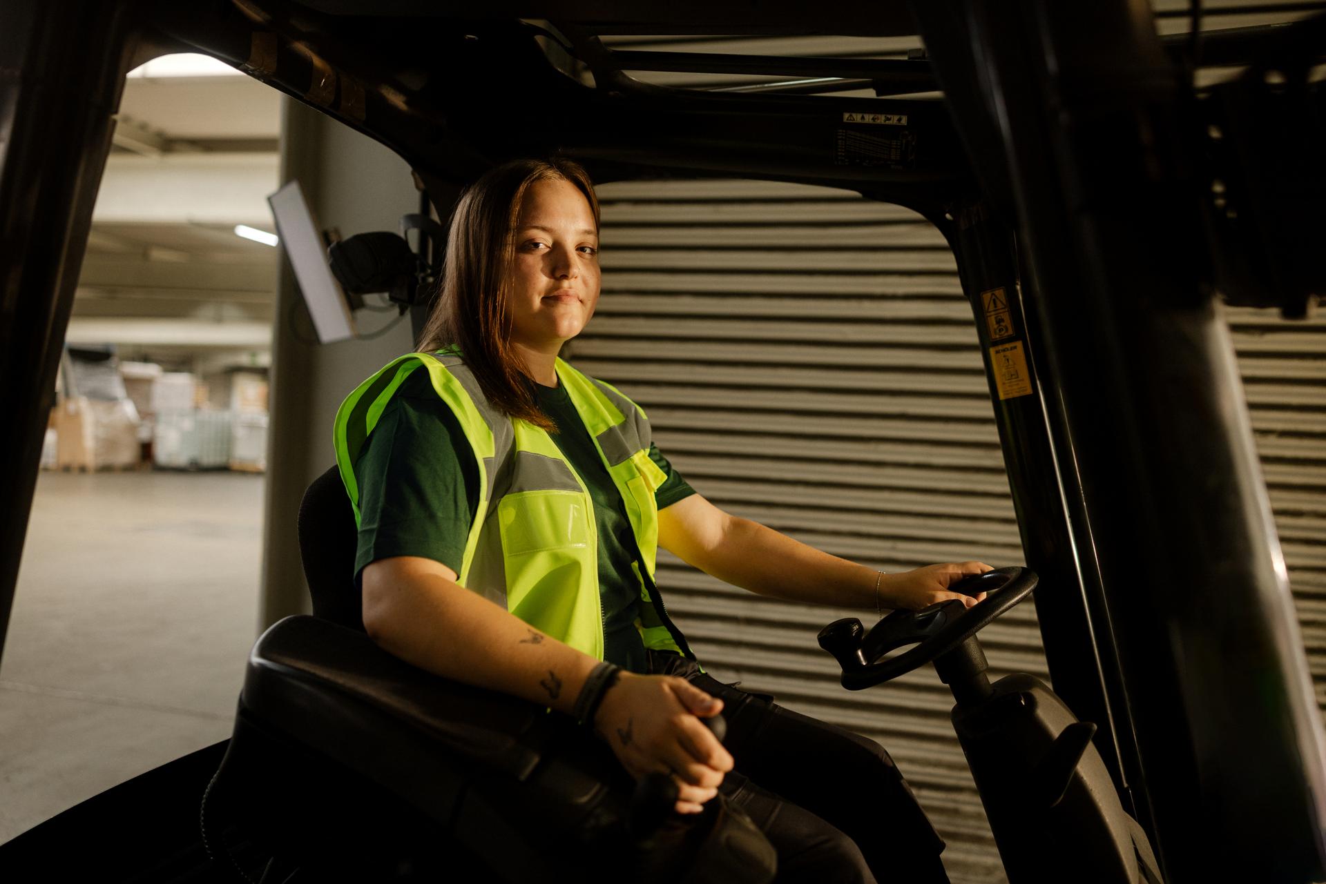 Une personne en tenue de travail bicolore se tient à côté d’un grand chariot en métal rempli de nombreuses caisses de transport empilées. Une main tient la poignée jaune du chariot. La scène se déroule dans un espace intérieur clair avec de grandes fenêtres et de longues ombres grâce à la lumière du soleil latérale.