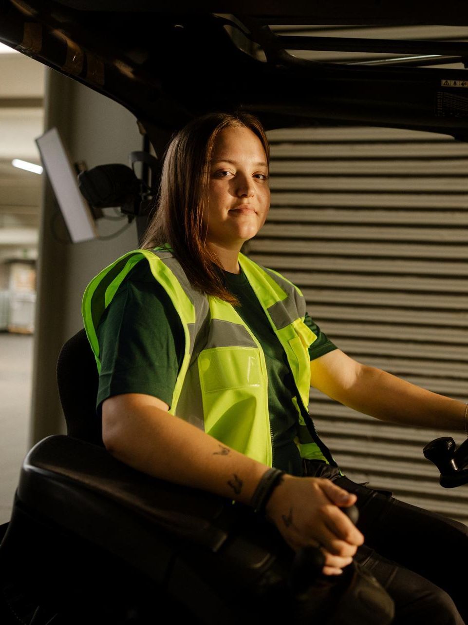 Une personne en tenue de travail bicolore se tient à côté d’un grand chariot en métal rempli de nombreuses caisses de transport empilées. Une main tient la poignée jaune du chariot. La scène se déroule dans un espace intérieur clair avec de grandes fenêtres et de longues ombres grâce à la lumière du soleil latérale.