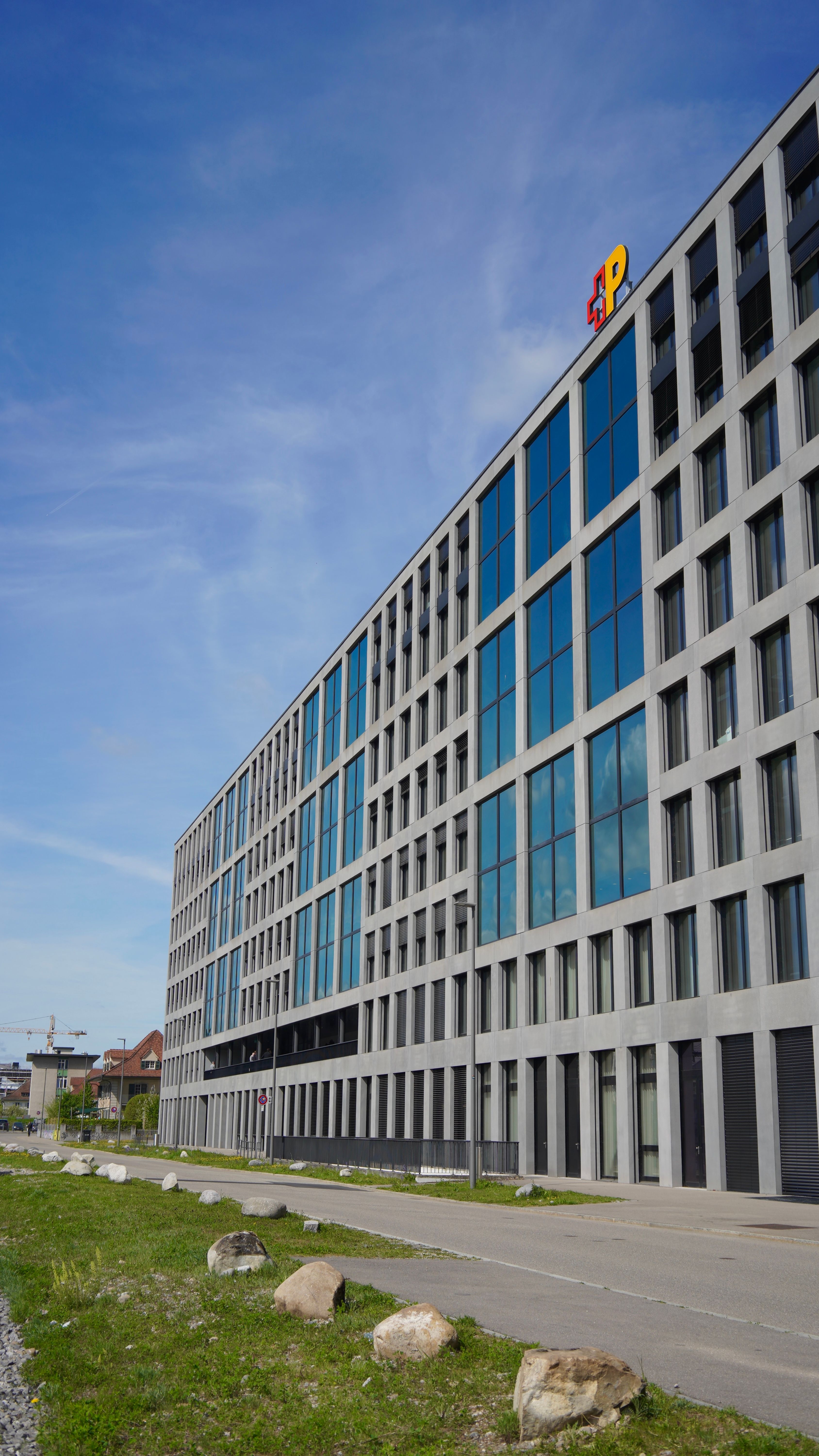 Modernes Bürogebäude mit Glasfassade und Logo der Schweizerischen Post auf dem Dach vor blauem Himmel.