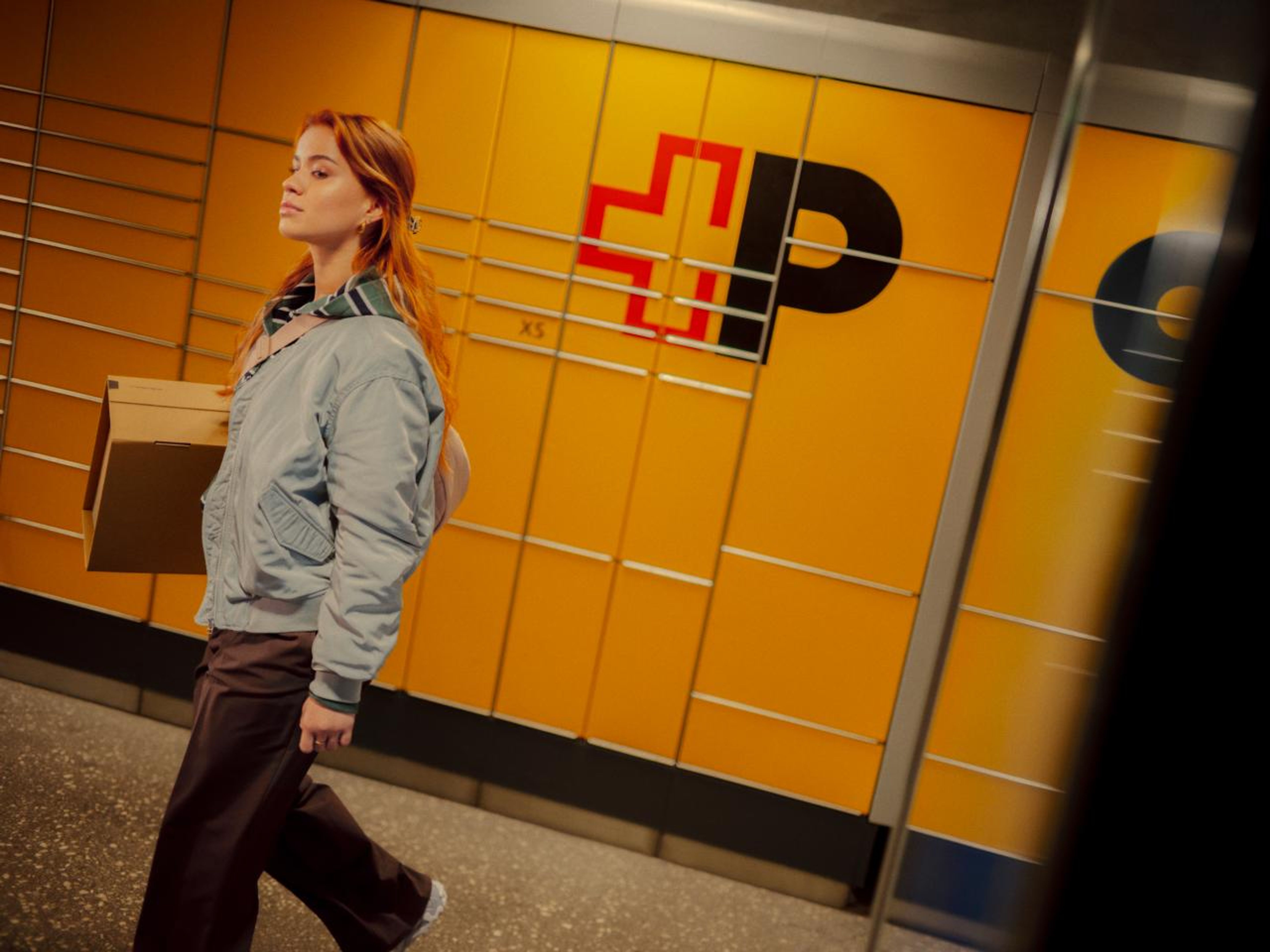 A person carries a parcel and walks past a yellow My Post 24 P.O. Box facility. The facility features the Swiss Post logo and is located in a modern, clean interior. The person looks relaxed and the room is well lit.