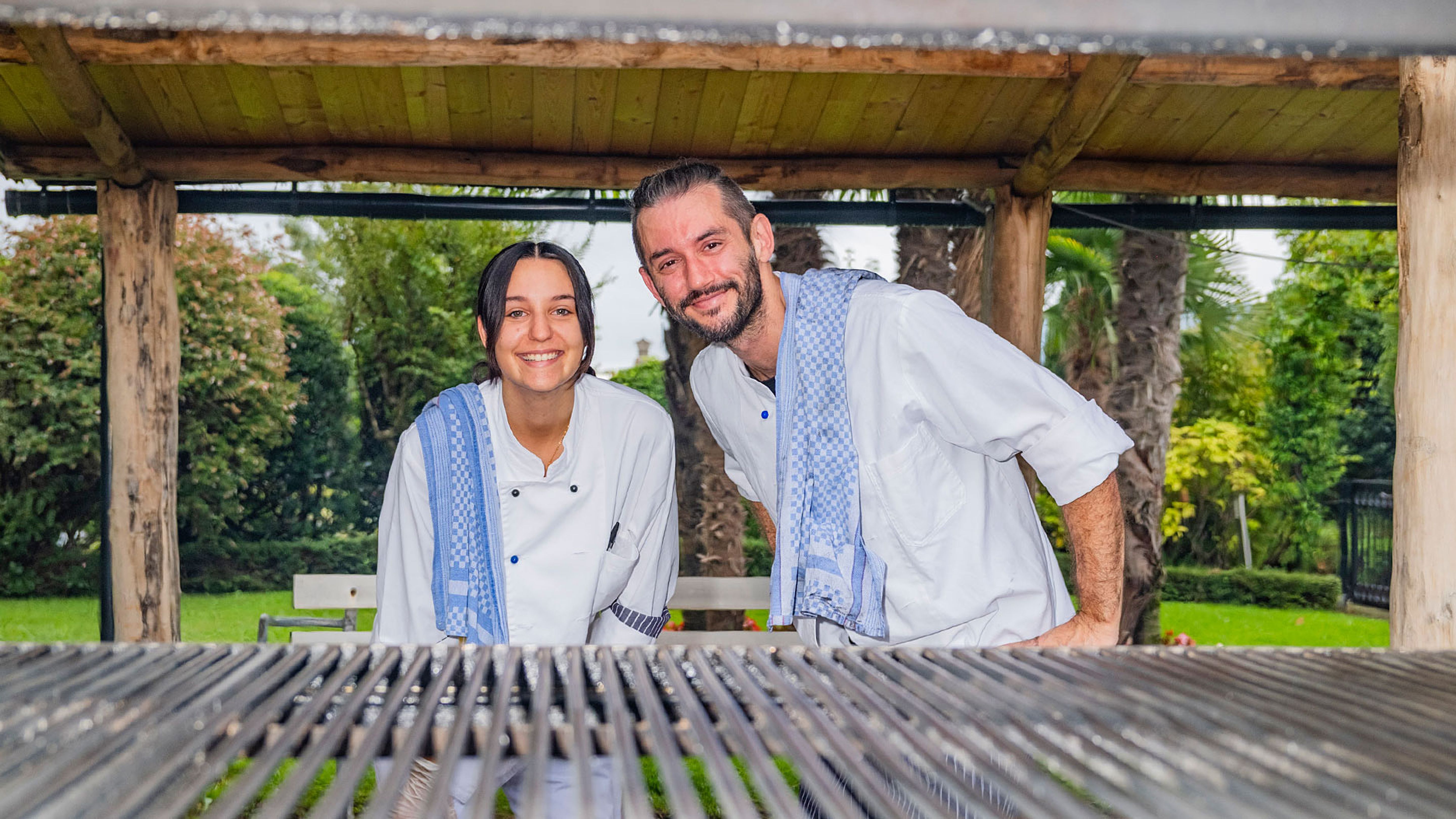 Deux personnes portant des vestes de cuisine blanches et des chiffons bleus dans une zone de jardin couverte de bois avec grille de barbecue.