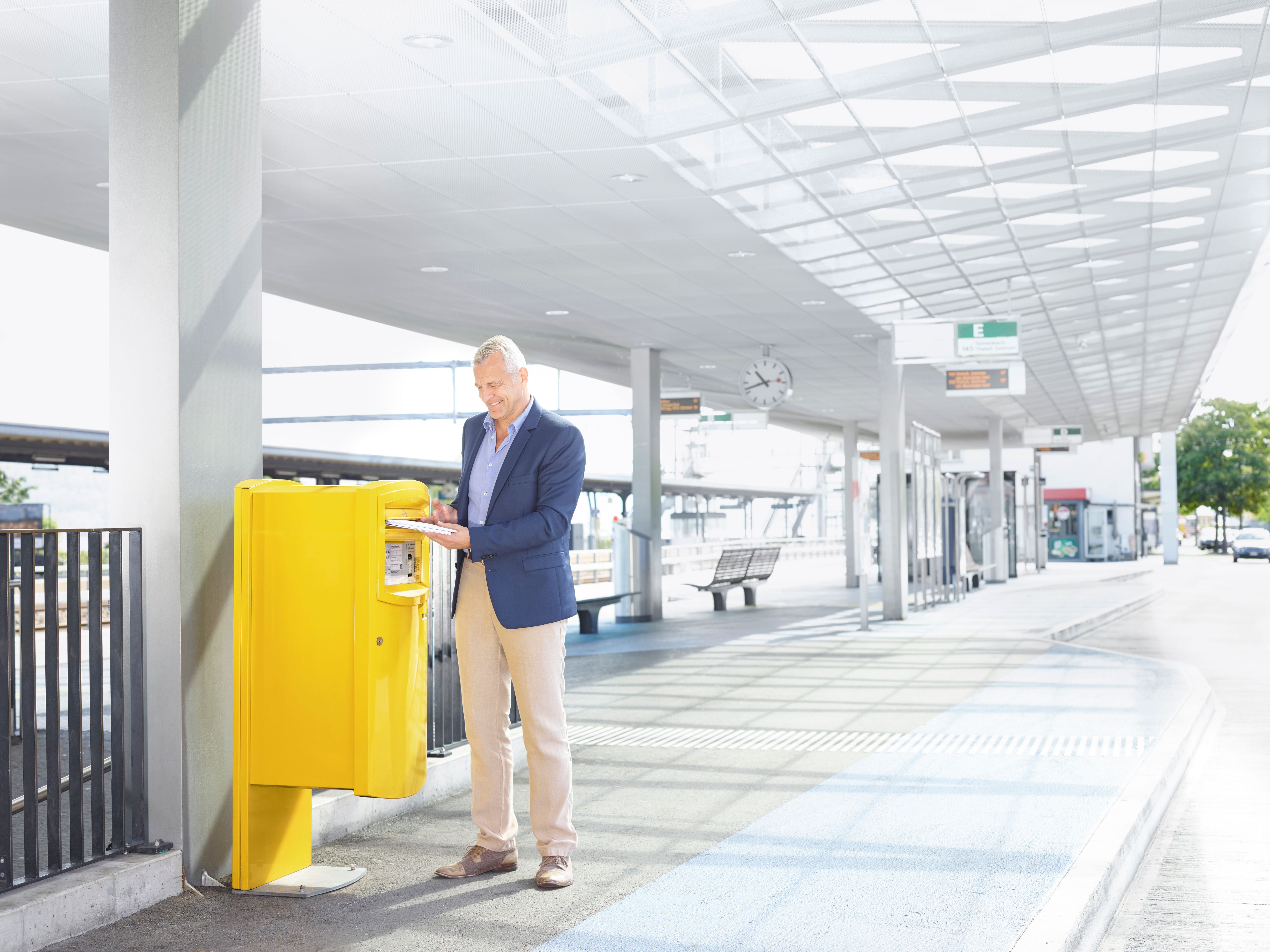 Mann wirft Brief in gelben Briefkasten an einem Bahnhof mit Uhr und Sitzbänken