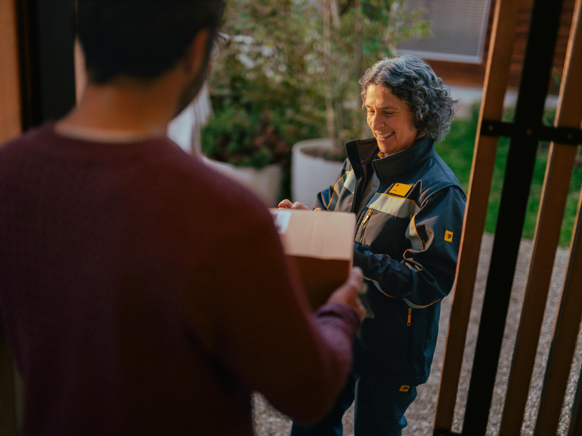 Une personne portant un uniforme de livraison remet un colis à une autre personne située au premier plan. L’échange a lieu devant une porte d’entrée, avec des plantes et un élément en bois en arrière-plan.