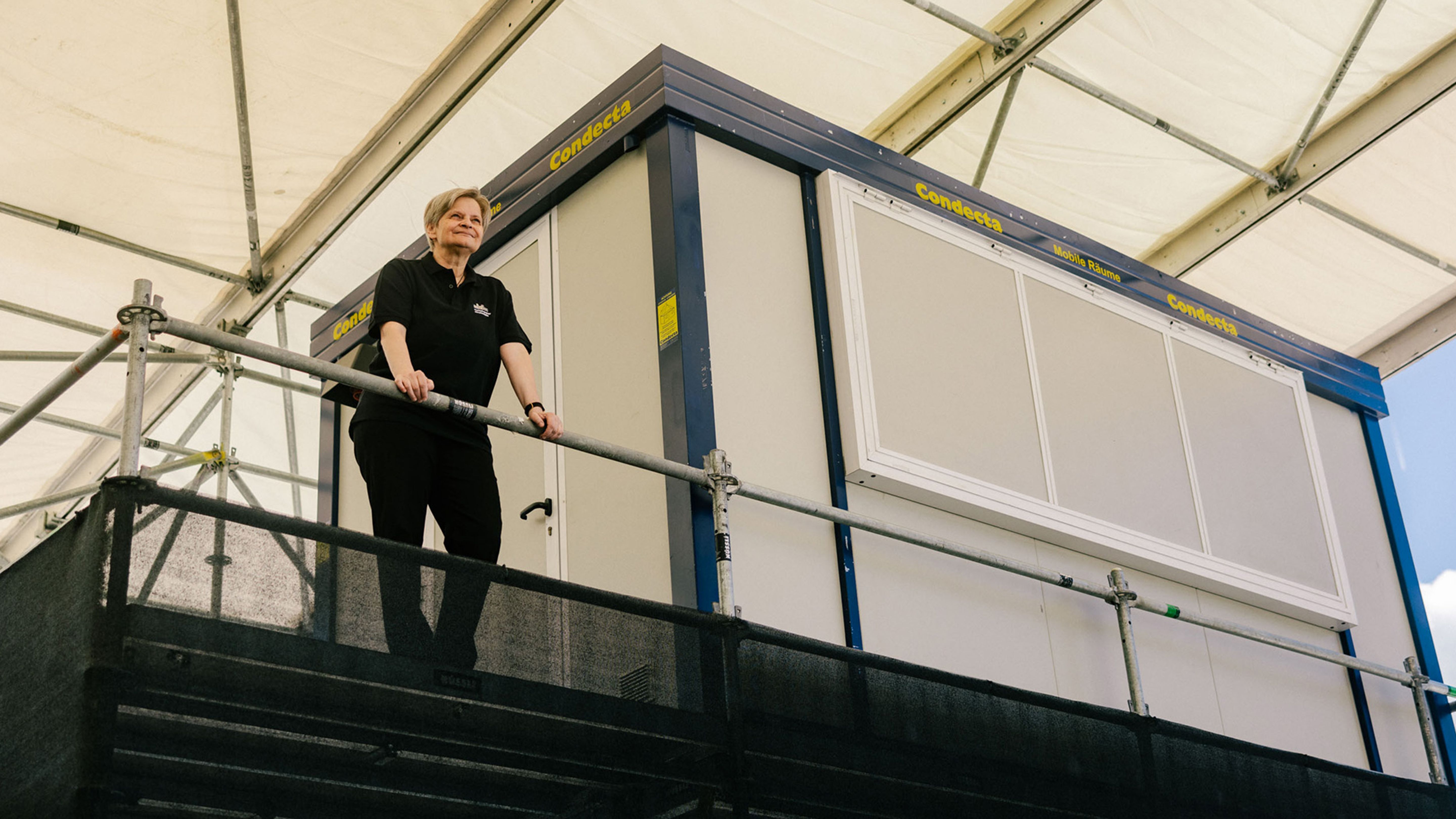 Eine Person steht auf einem Gerüst vor einem modularen Gebäude unter einem Zelt.