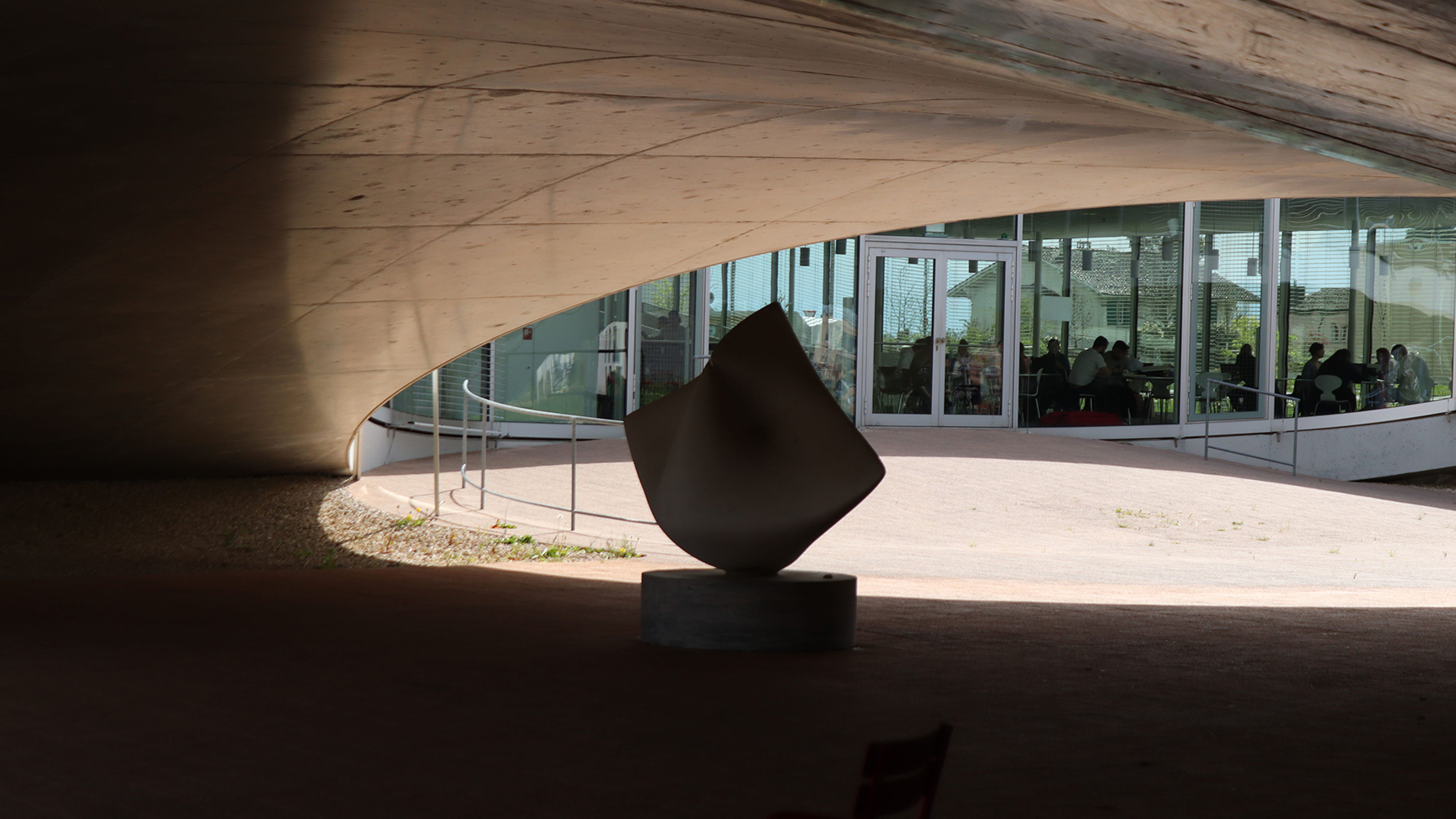 Une sculpture se trouve sous un plafond en béton incurvé, avec des vitraux et des sièges en arrière-plan.