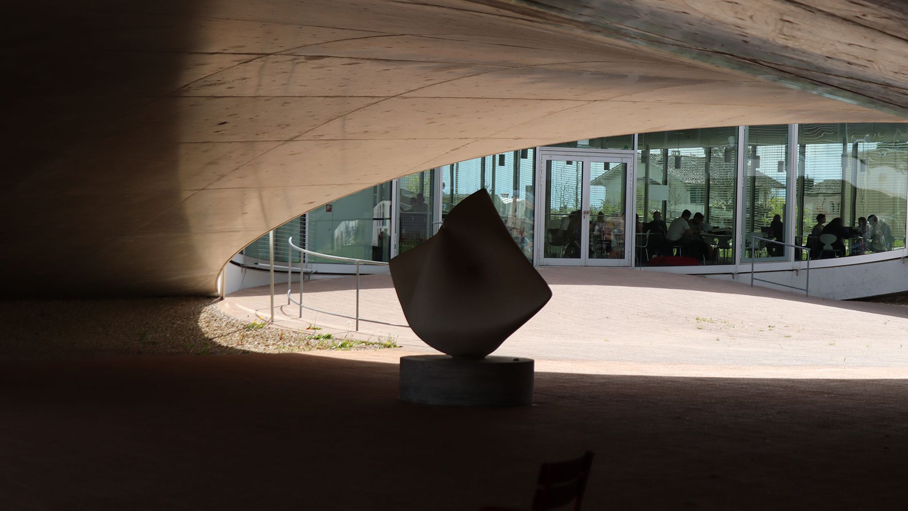 Une sculpture se trouve sous un plafond en béton incurvé, avec des vitraux et des sièges en arrière-plan.