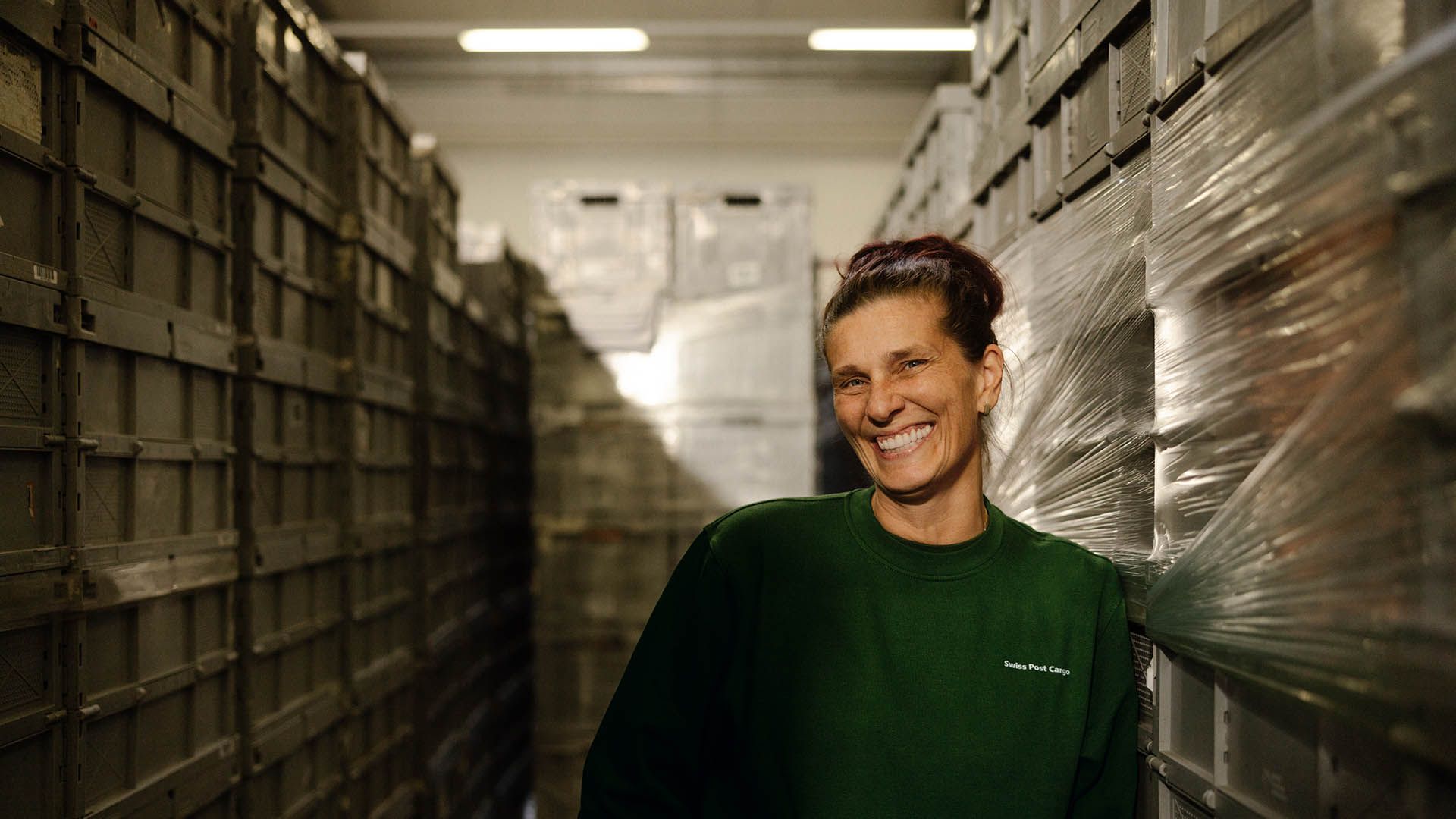 A logistics employee from Swiss Post Cargo is smiling while standing between stacked transport crates in a warehouse.