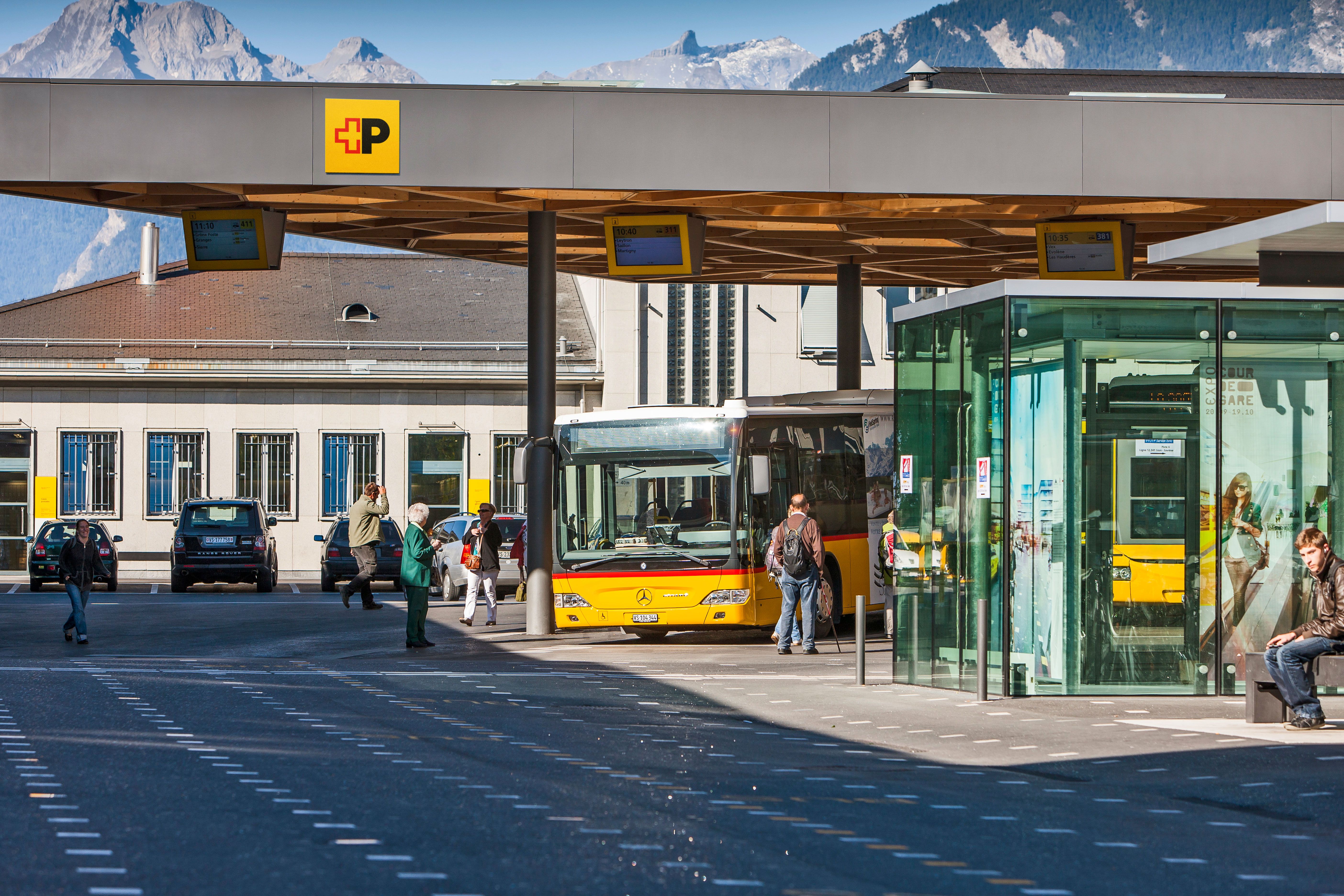 Gelb-roter Postbus an Bushaltestelle mit wartenden Personen und Bergen im Hintergrund