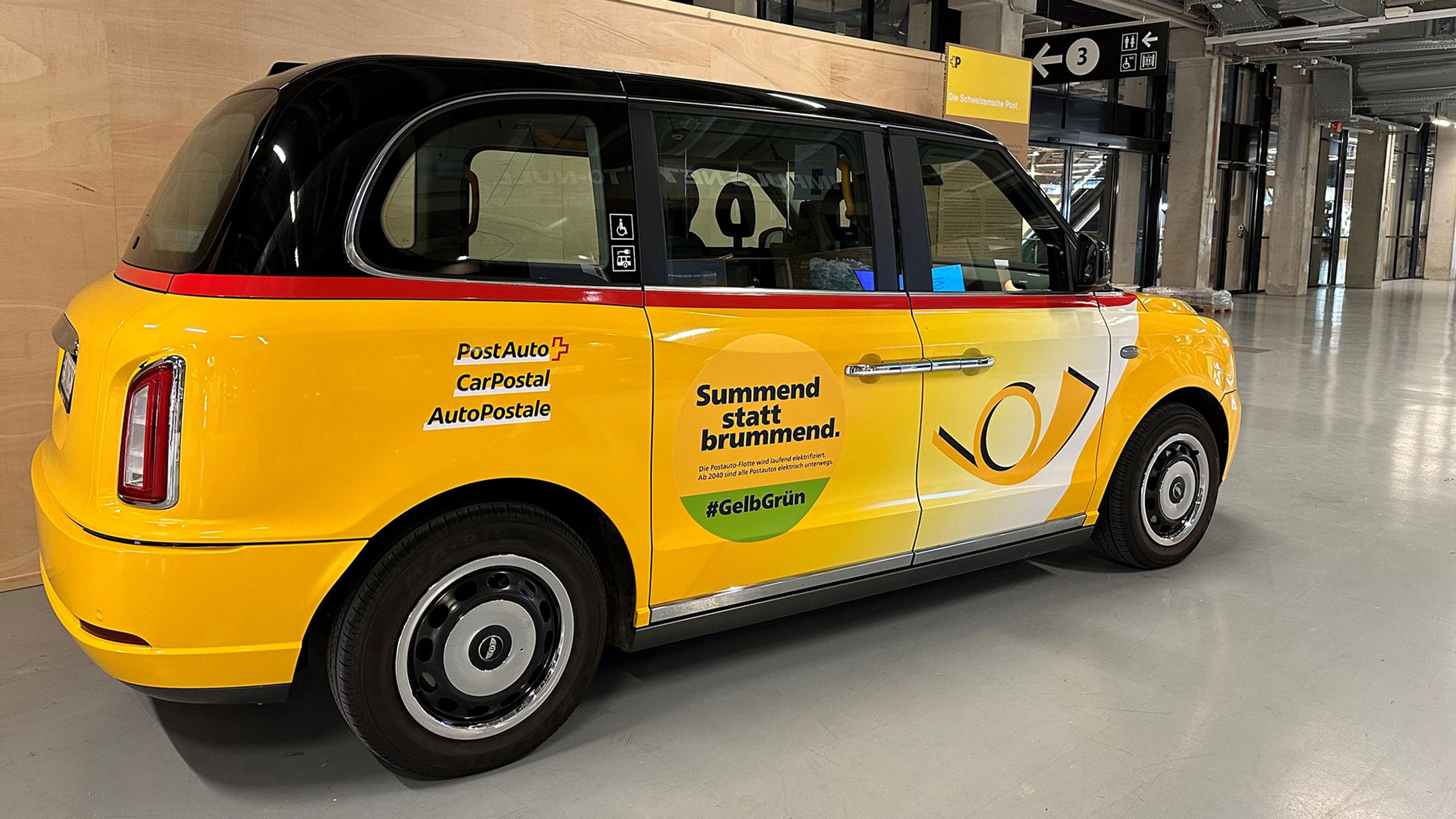 Ein gelbes Fahrzeug mit dem Logo der Post und Elektromobilitätsaufklebern steht im Innenbereich.
