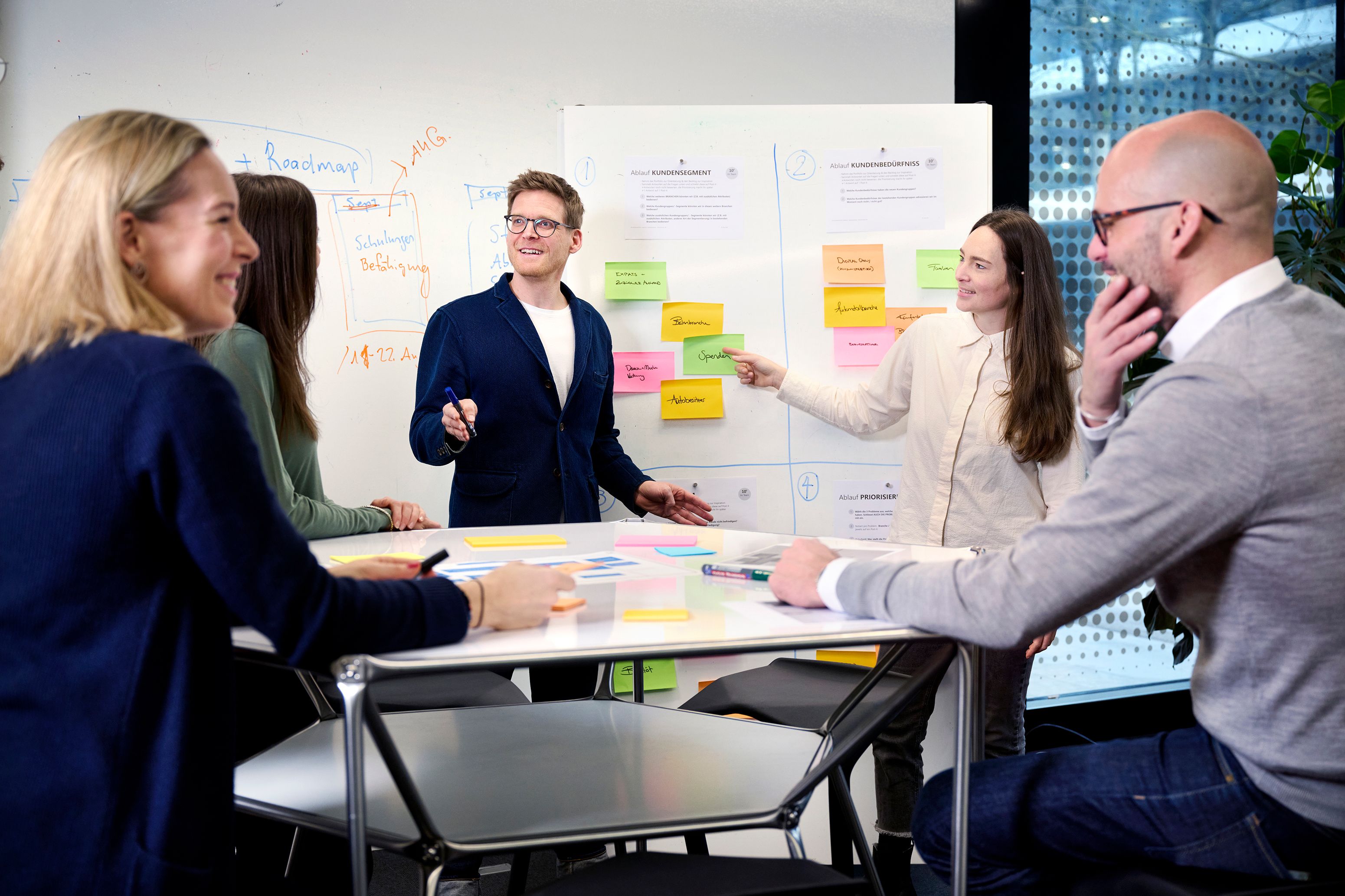 Eine Gruppe von Personen in einem Büro diskutiert vor einem mit Notizzetteln bedeckten Whiteboard.