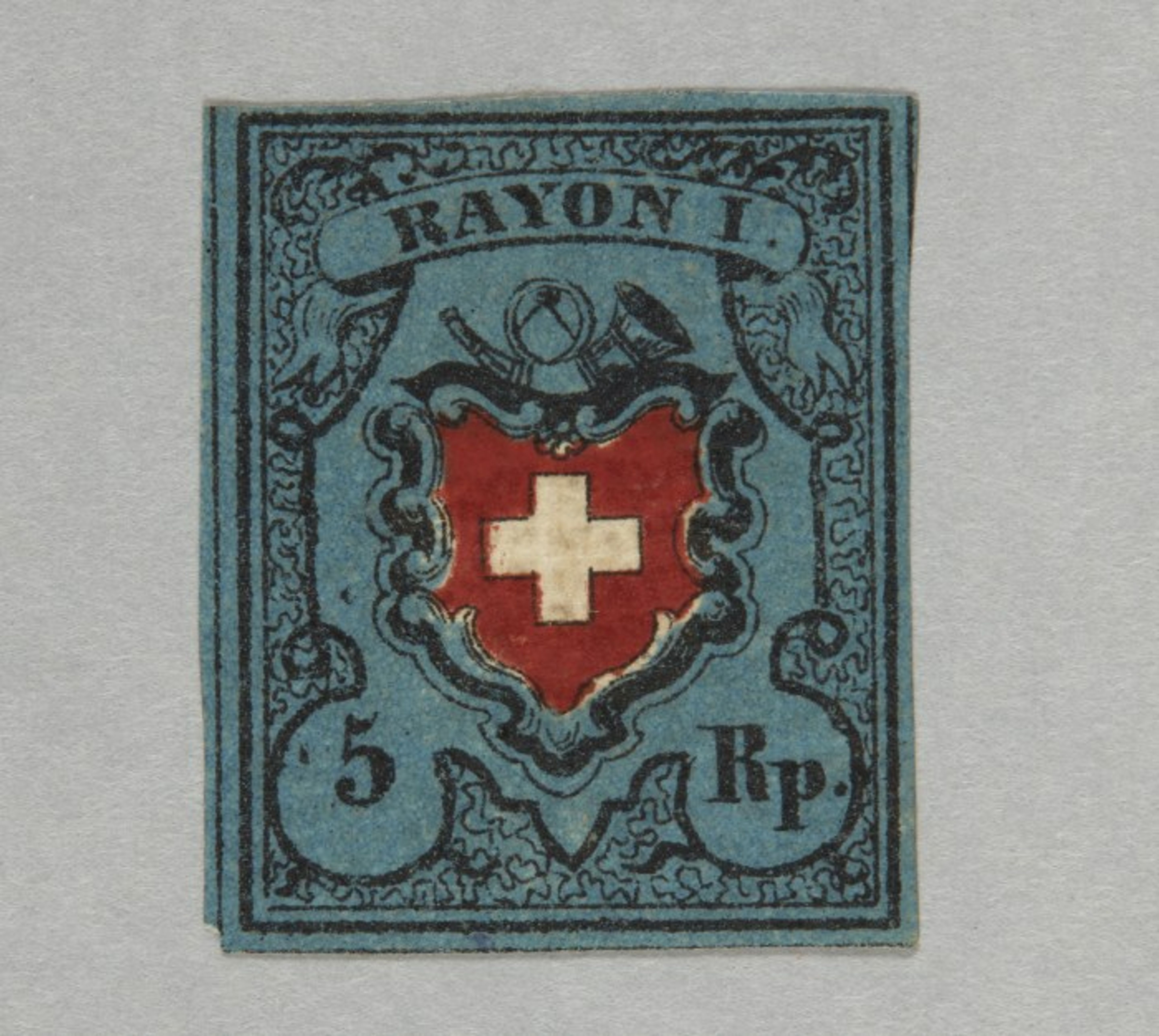 Timbre postal historique suisse avec l’inscription ‘Rayon I’. Au centre, un blason rouge avec une croix blanche, entouré d’un cadre décoratif bleu. En haut, on voit un cor postal et une trompette. En bas figure la valeur ‘5 Rp.’.