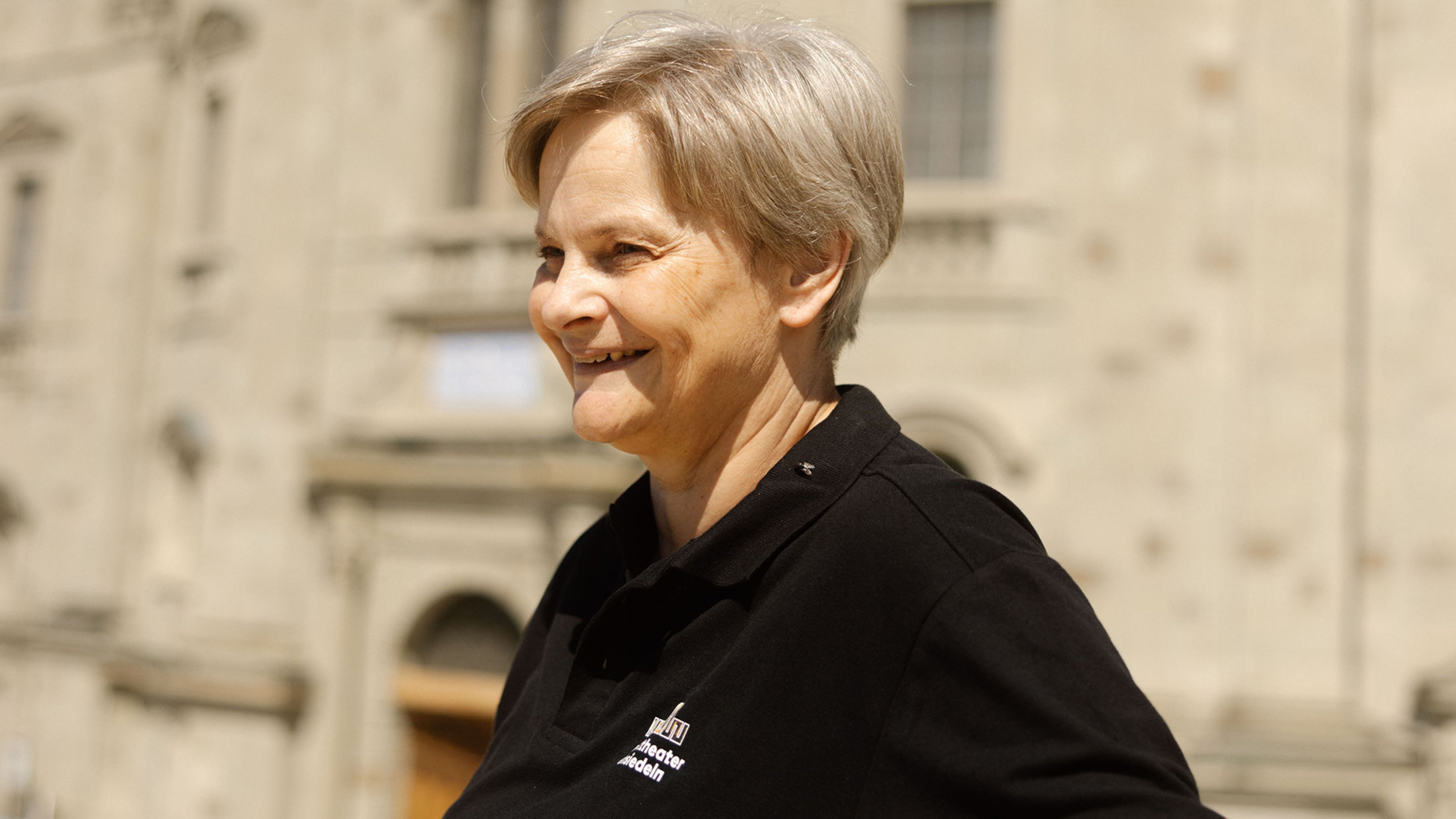 Eine lächelnde Frau im schwarzen Poloshirt steht vor einer historischen Fassade.