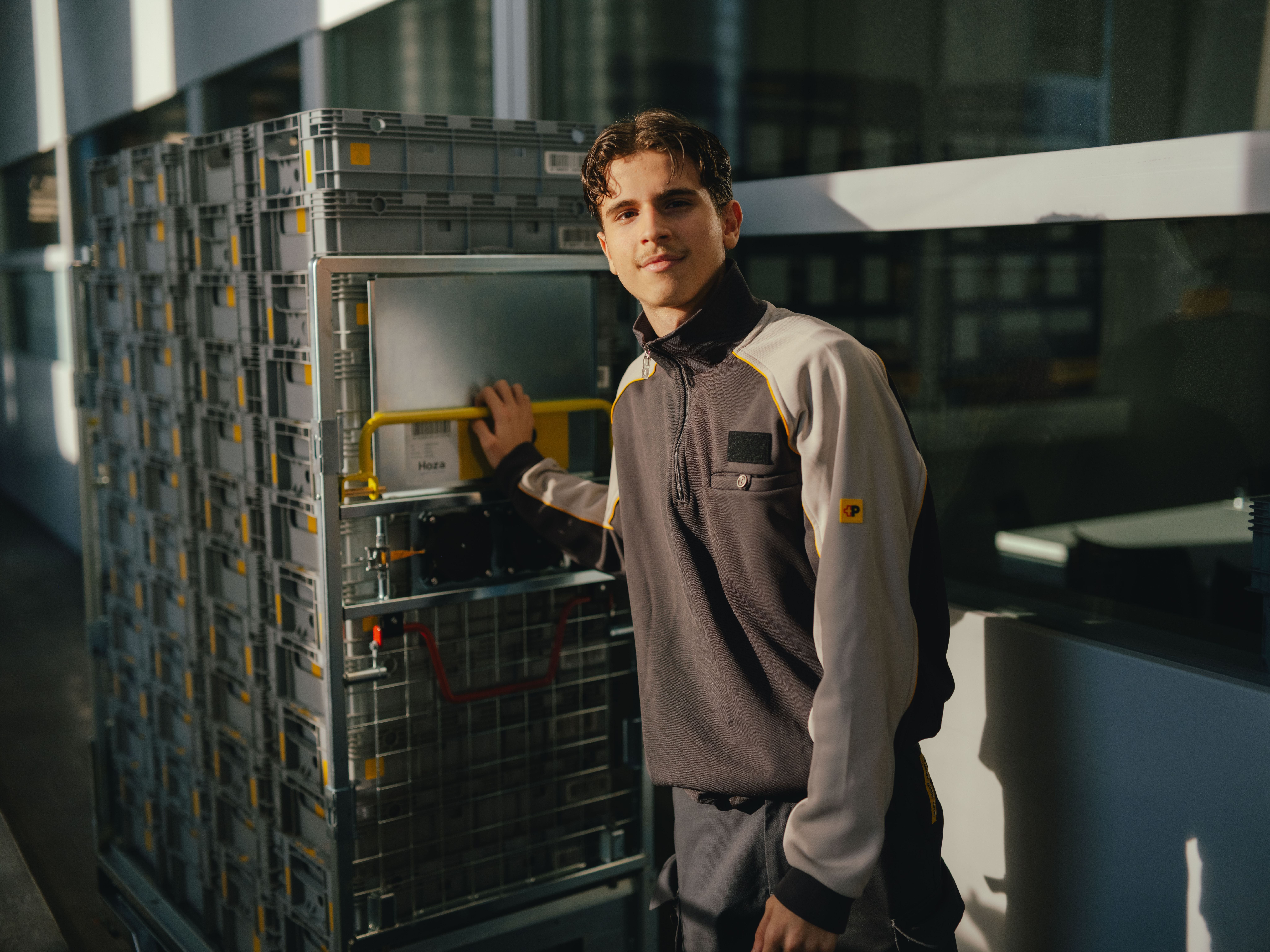 A person in work clothes is standing next to a high metal trolley with several stacked transport crates. One hand is holding the yellow handle of the trolley. The scene is located in an interior area with large windows that let sunlight in.